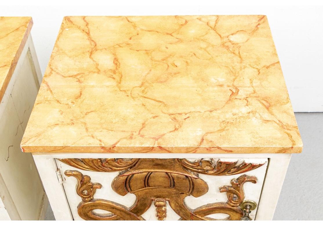 Square stands raised on tapering square legs in white paint with faux marble painted tops in red and yellow. The case with a hinged door over a lower drawer, with a large raised carved gilt foliate scrolled motif top to bottom. Both with fine flower