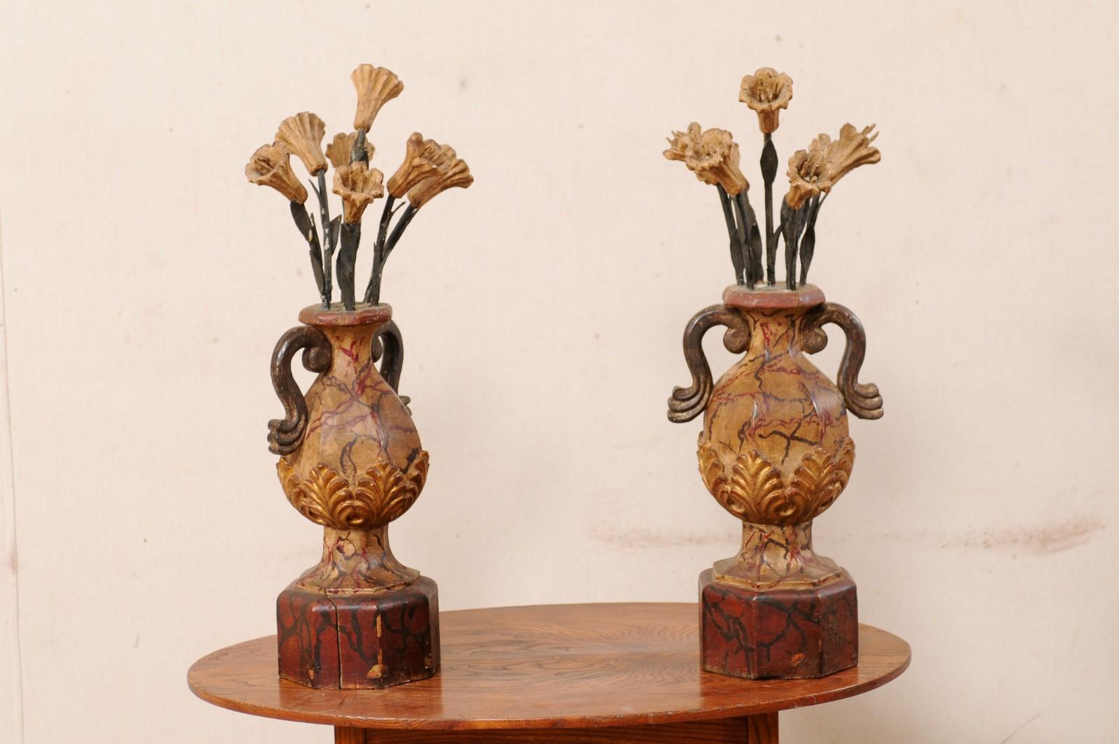 An Italian pair of carved wood urns from the early 20th century. This antique pair of decorations from Italy each feature a hand-carved, two-handled urn, which is raised upon a pedestal base, with a floral bouquet springing upward out from it's top.