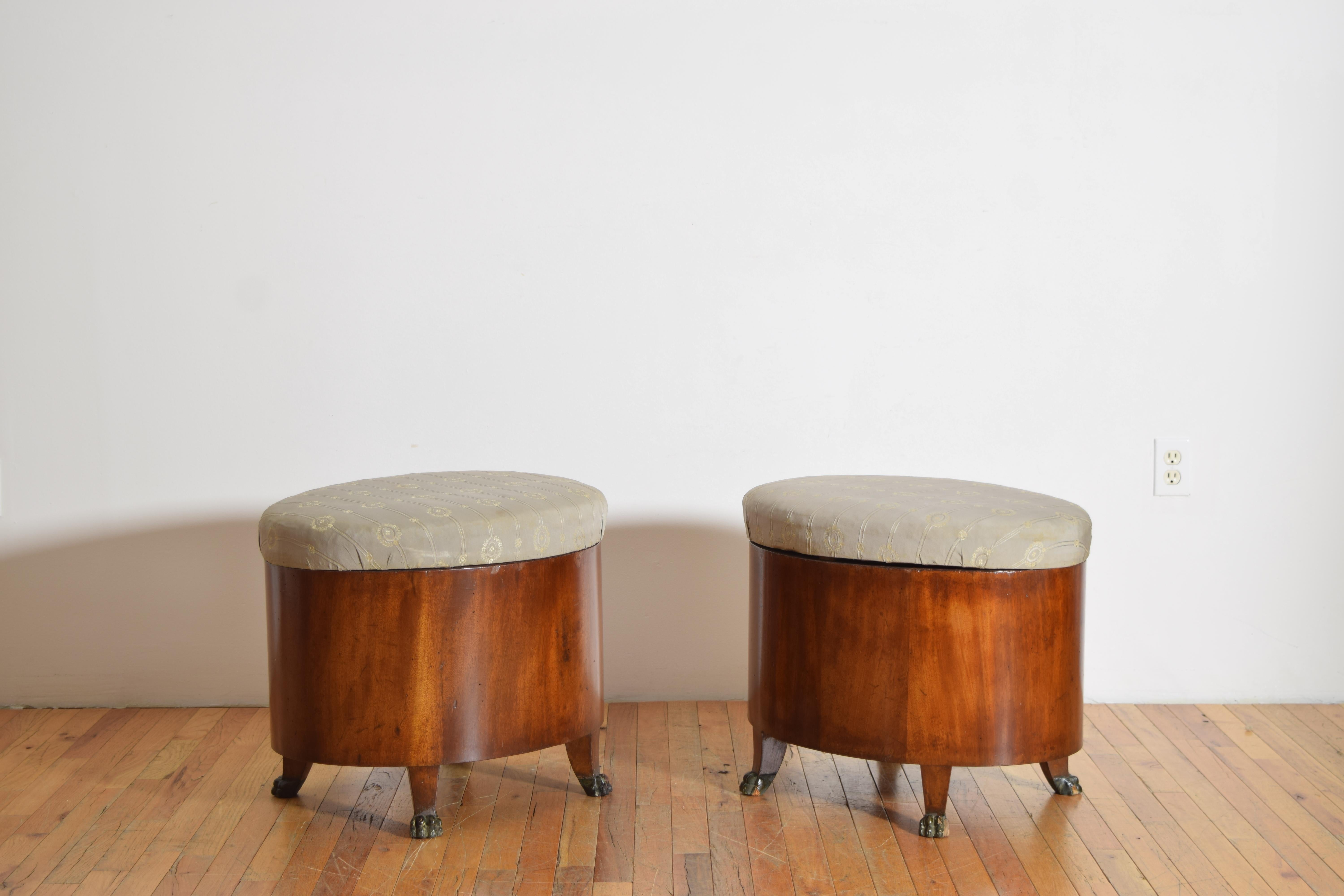 each bench having a removable upholstered oval top resting upon a conforming case with a large storage well within, the cases raised on small splayed legs terminating in carved and ebonized paw feet