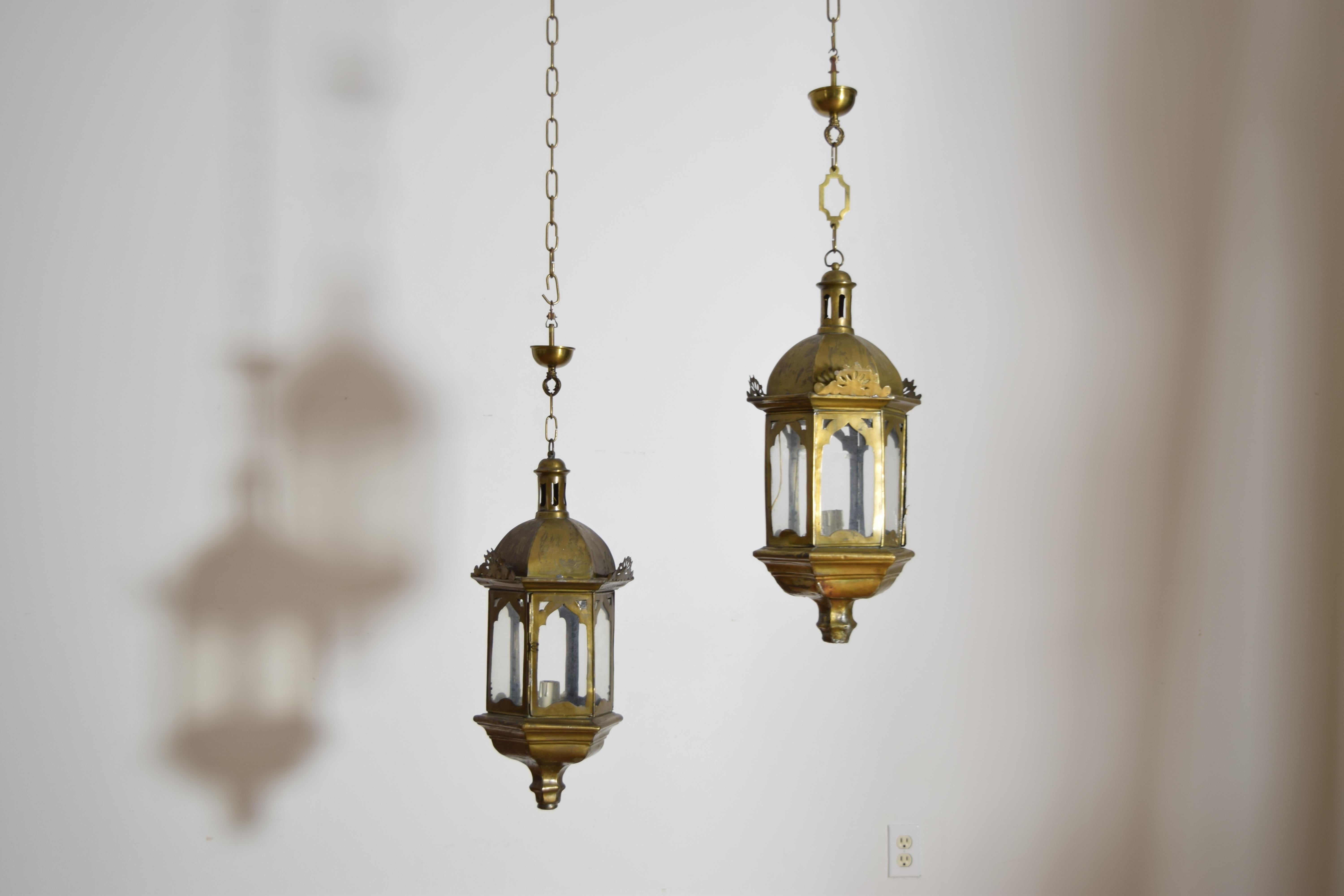 these elaborately constructed lanterns are hexagonal and have a number of decorative features with graduated bases, brass overlay in patterns on the glass sides, 3 sides with decorative pediments, dome-fork hexagonal tops, and turret-form finials