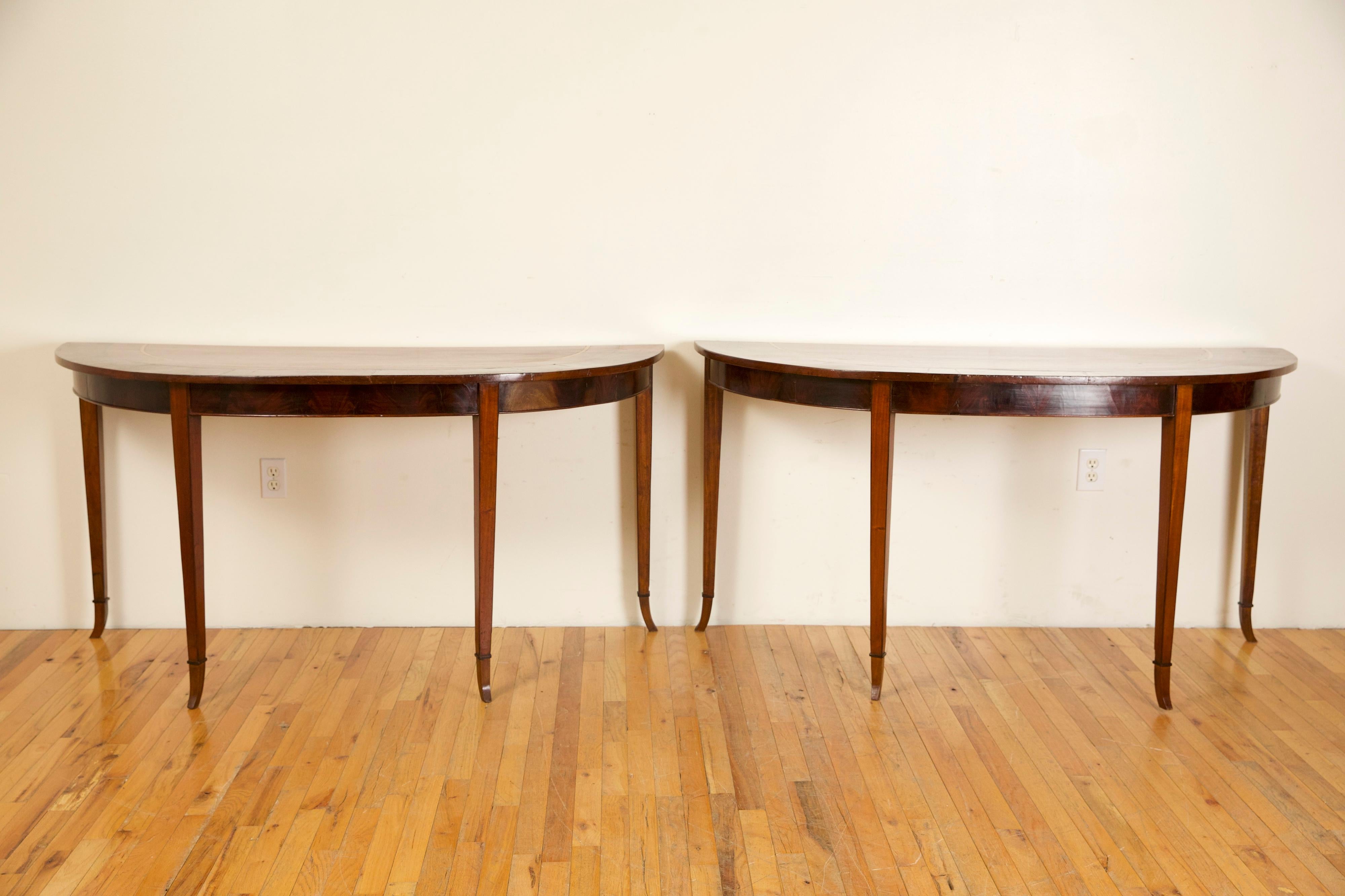 Likely from Naples, Italy, these wide and shallow consoles are made entire from mahogany with light walnut inlaid banding, demi-lune in shape with conforming aprons and raised on square tapering legs ending in stylized curved feet