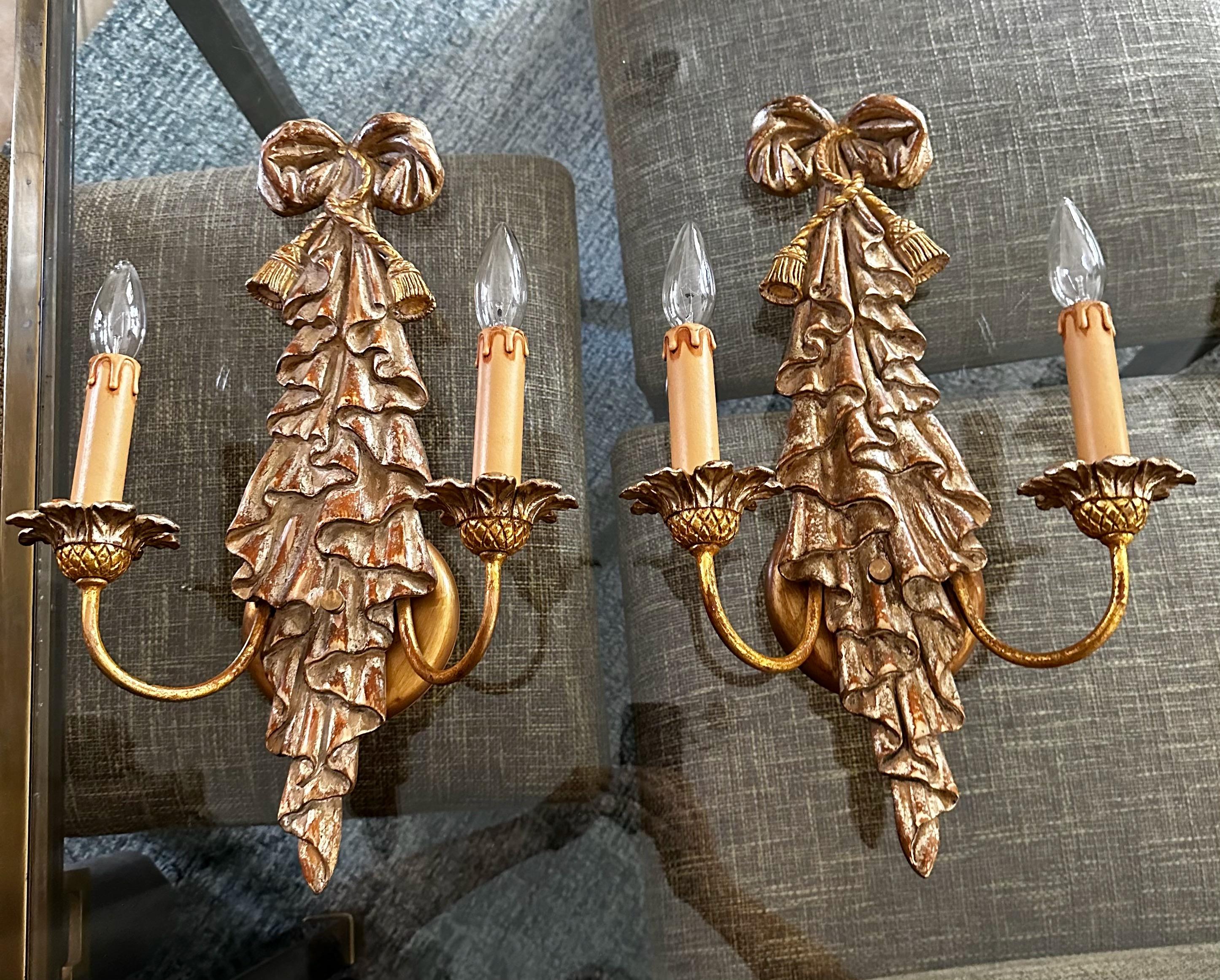 Paire d'appliques murales à deux bras de style néoclassique en bois doré sculpté, avec des reflets dorés et argentés. Le design des appliques comprend un ruban un motif drapé. Chaque applique utilise deux ampoules candélabres à culot 