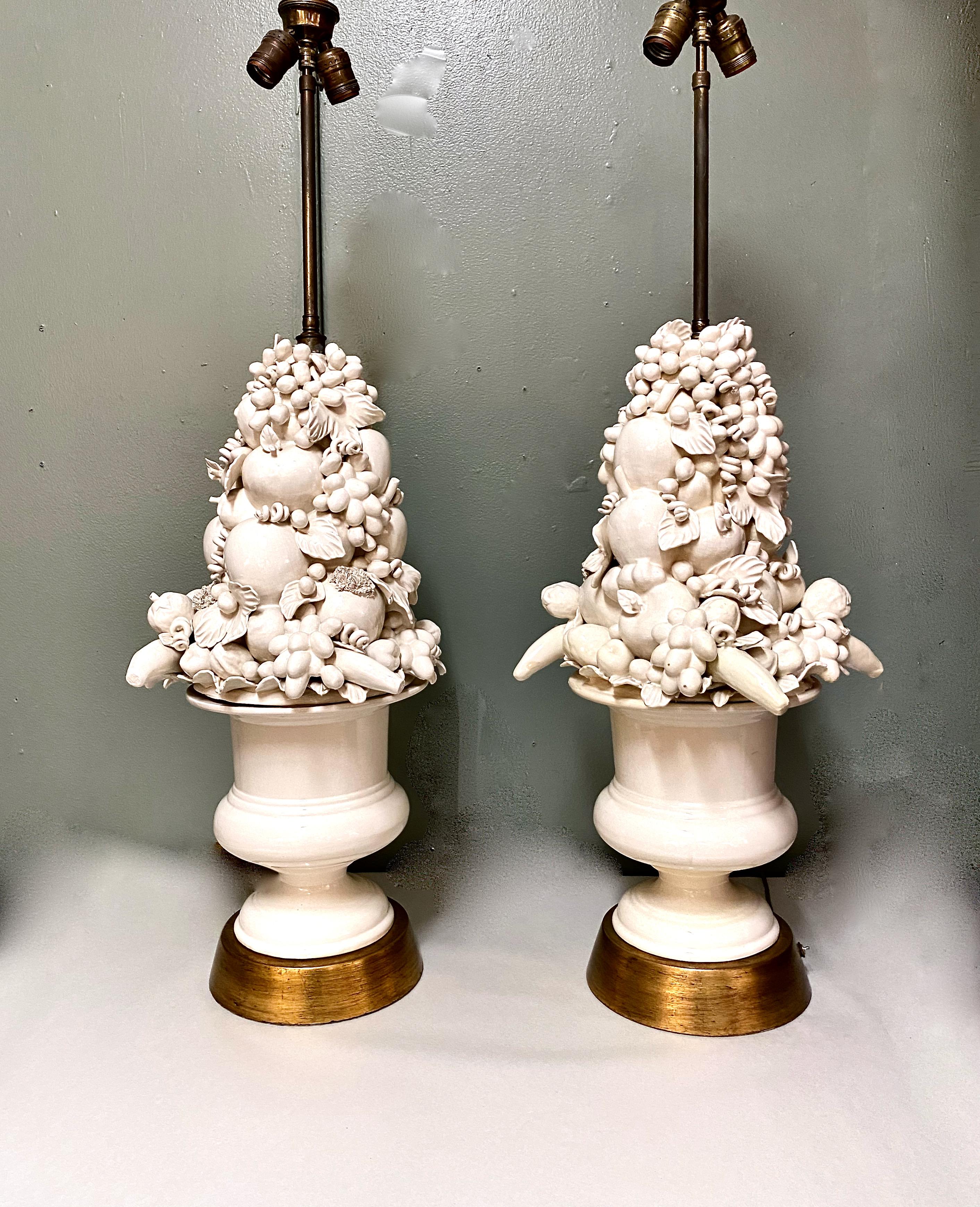 This is a stunning pair of unusual mid-20th century Italian pottery lamps composed of a neoclassical style urn heaped with various fruits. The white body of the lamps is highlighted by a substantial gold leafed plinth. The double cluster electrical