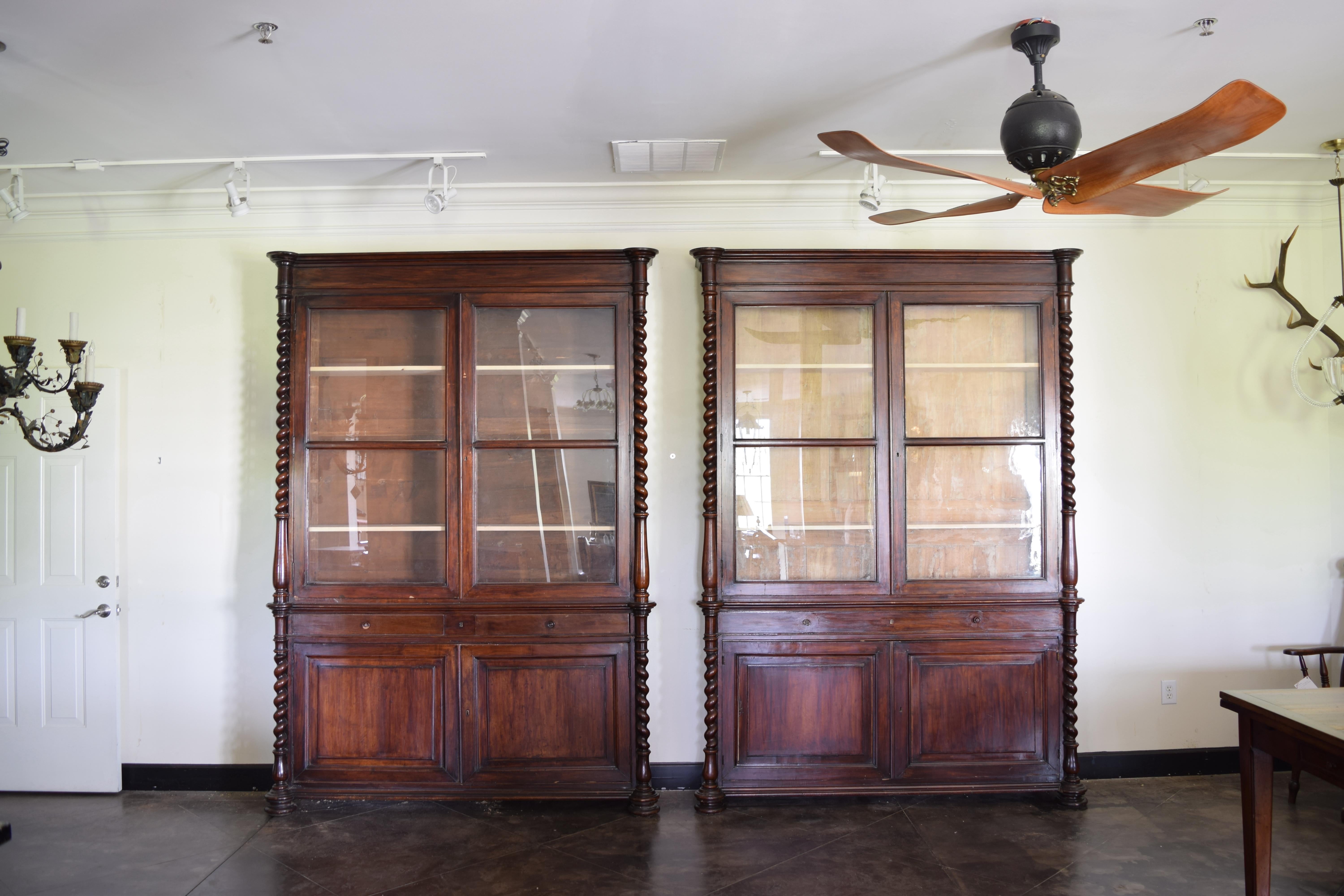 tall and shallow these impressive cabinets are of two piece construction, each with turned columns on upper and lower sections, the tops with glass fronts, the bottoms one with two drawers the other with faux drawers, both sections with interior