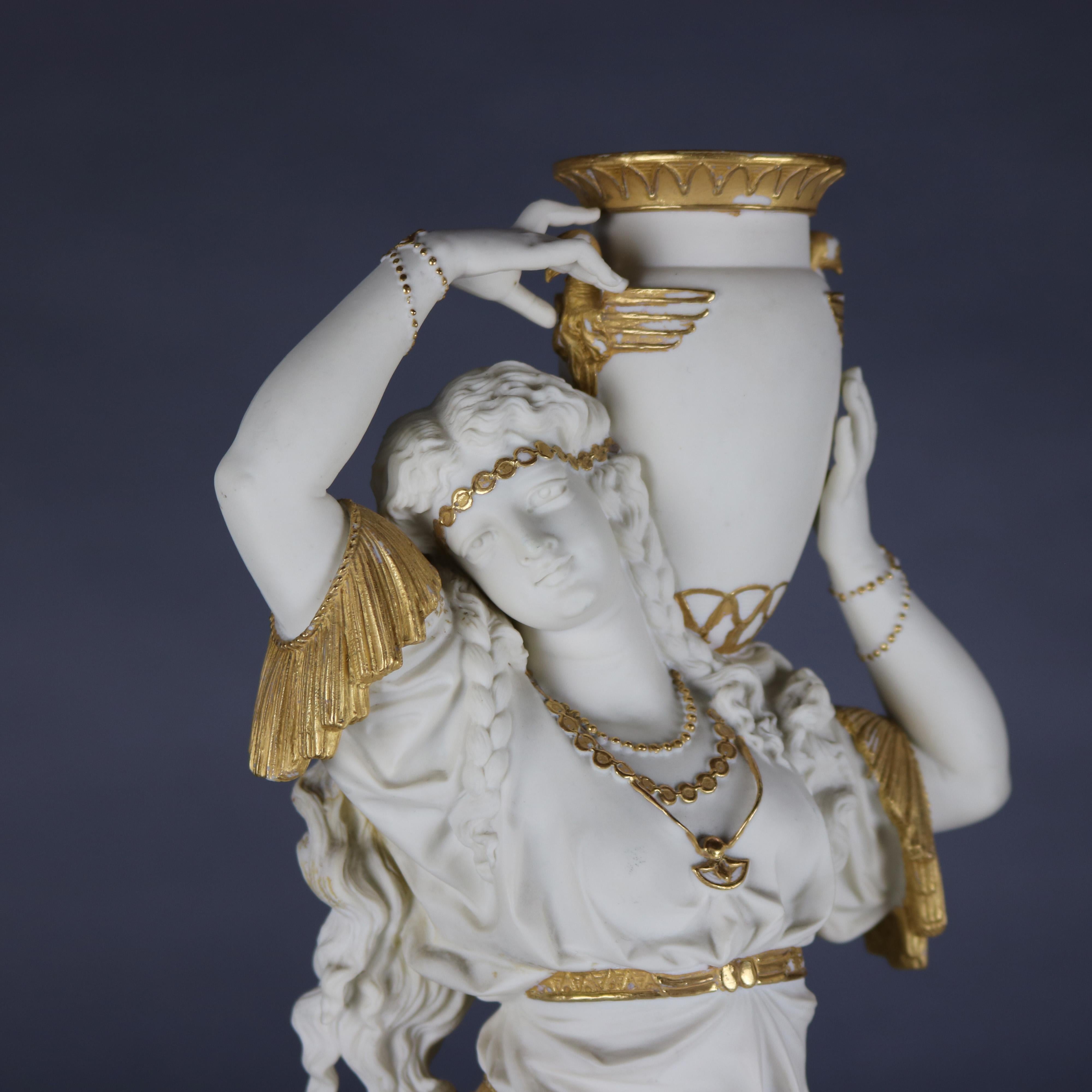 An antique pair of large Egyptian Revival figural statues by J. E. Caldwell offer bisque porcelain ladies carrying water vessels in countryside setting and raised on footed plinths, gilt highlights throughout, on base 