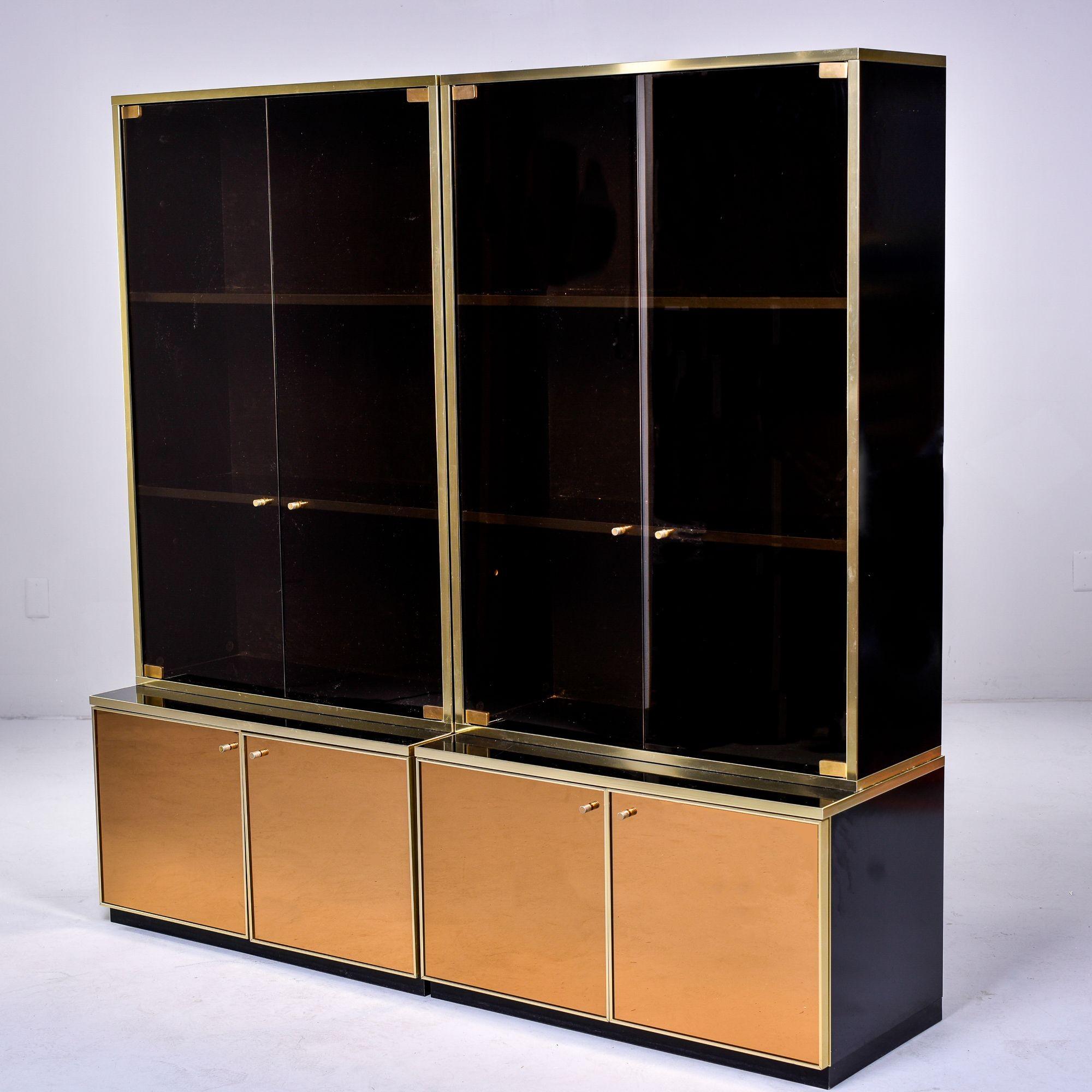 Found in France, this pair of circa 1980s two piece shelf cabinets have black glass top sections and a mirrored copper colored base - all are trimmed in brass. Top sections each have two near-black glass hinged doors and two fixed internal shelves.