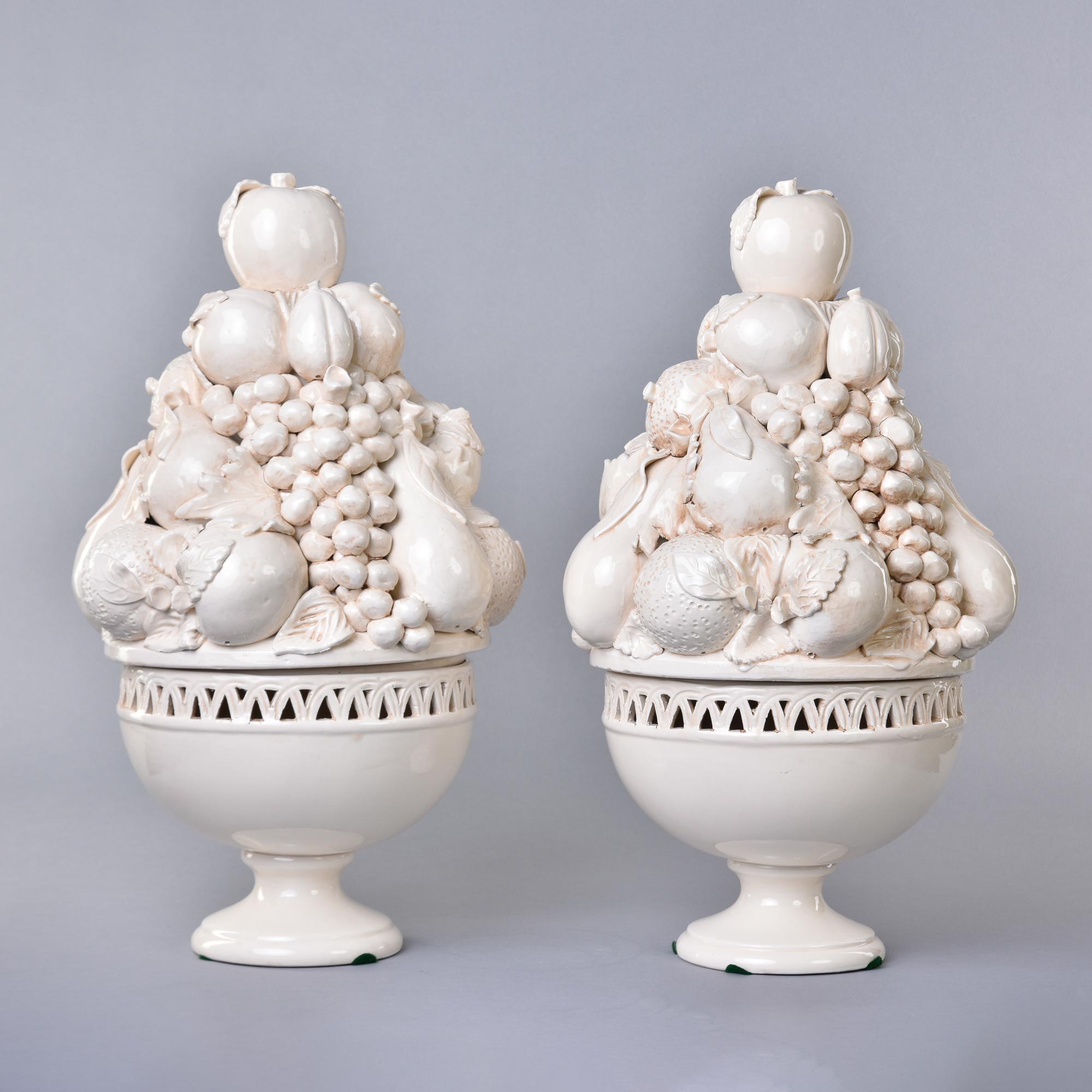 Found in Italy, this is a new pair of Italian-made lidded fruit garniture bowls in a creamy off-white glaze. Pedestal base with rim of bowl decorated with cut out border. Removable tops are well-rendered tall ceramic fruit displays depicting grapes,