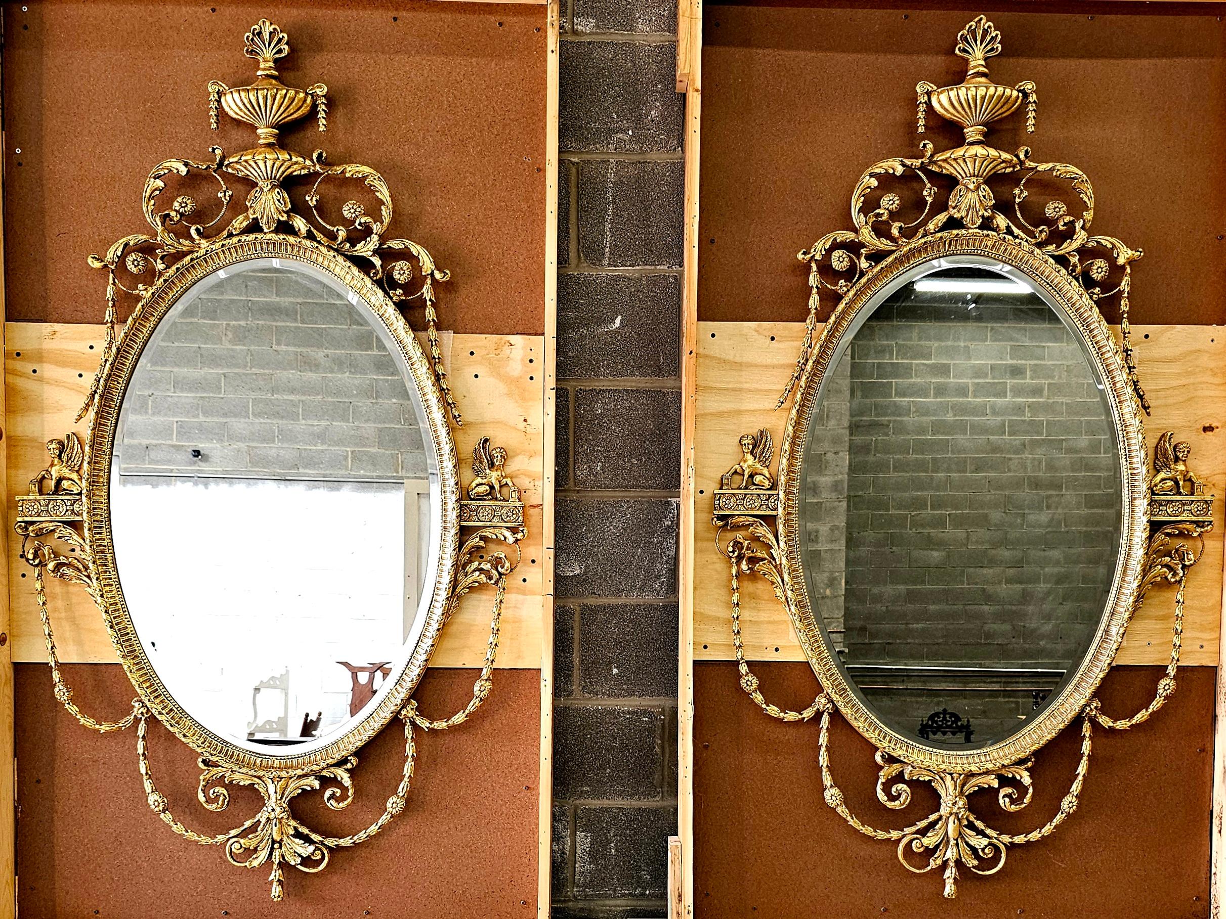 Beautiful large pair of Adam style carved giltwood oval wall mirrors. This a beautiful pair of beveled mirrors with double urns and foliated garland cresting measuring 35