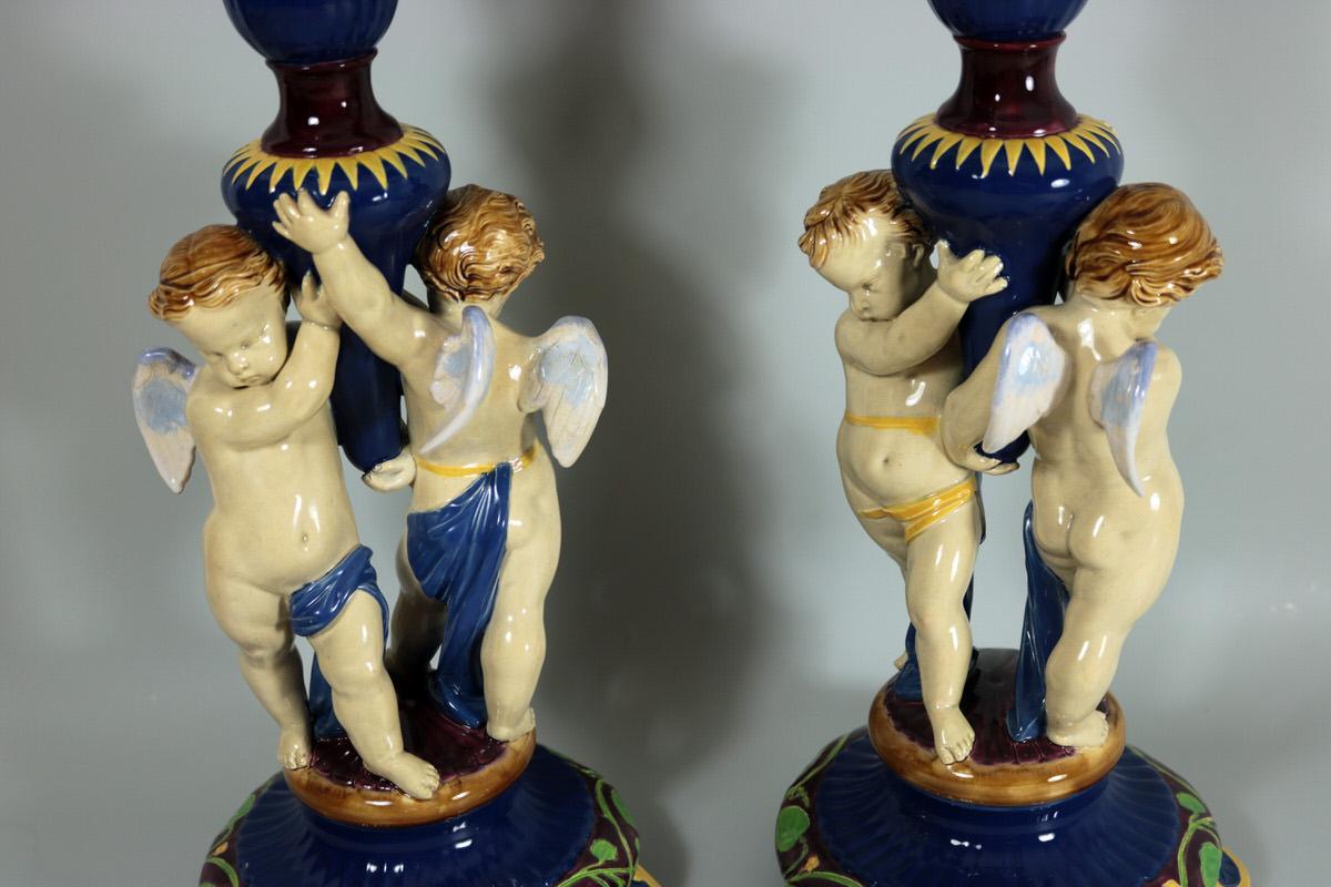 Pair of Minton Majolica vase figural which feature a pair of winged cherubs, supporting a large trumpet vase. Coloration: Minton-blue, cream, green, are predominant. The piece bears maker's marks for the Minton pottery. Marks include a factory