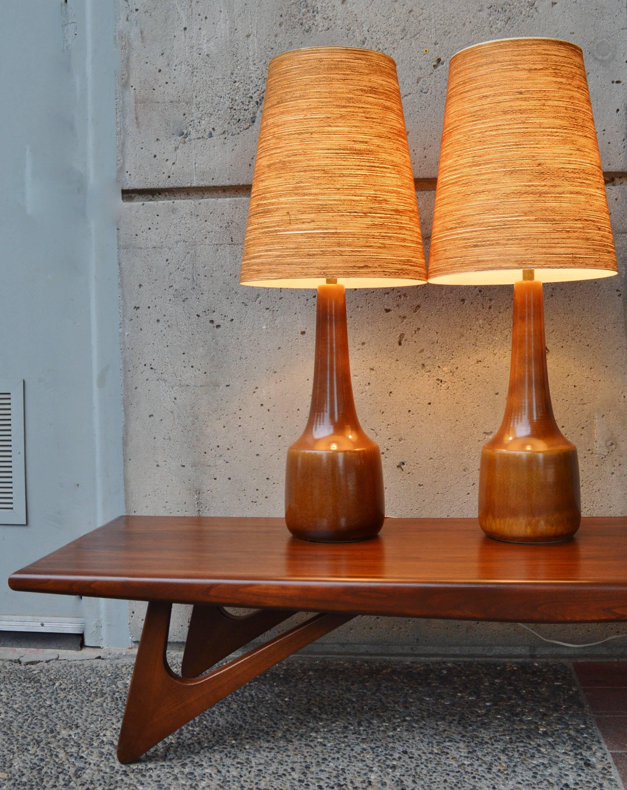 This stunning pair of tall ceramic lamps were handcrafted in the 1960s by Danish potters Lotte & Gunnar Bostlund. The subtle glazing variations in caramel earth tones is spectacular. Featuring their three-tier original brass finials and uniquely