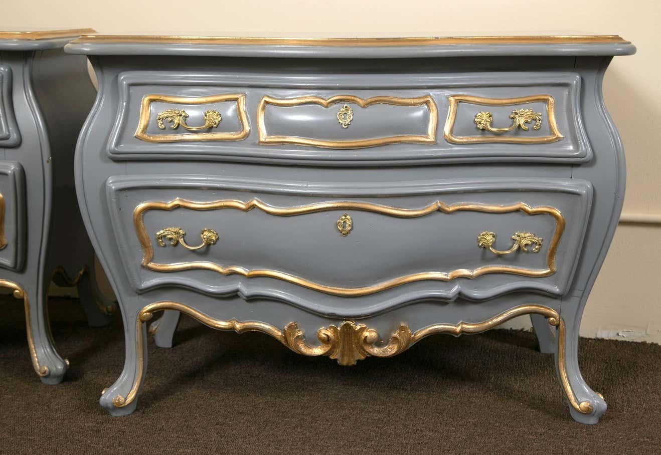 A fine Hollywood Regency pair of parcel paint and gilt decorated bombe commodes. These Louis XV style commodes or nightstands, each in an off gray color with gilt gold highlights. Bombe in form with two drawers having bronze pulls and key holes. The
