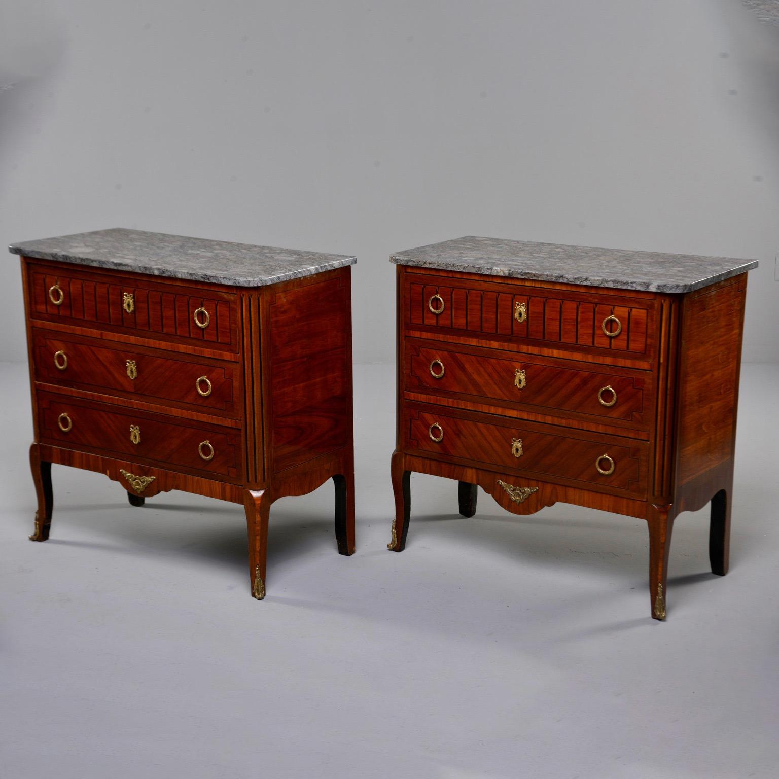 Found in Italy, this pair of French cabinets date from circa 1900. Each piece is mahogany with three functional drawers, wreath-style brass pull rings, ribbon-crested escutcheons, decorative brass mounts on the apron and feet, and dark gray marble