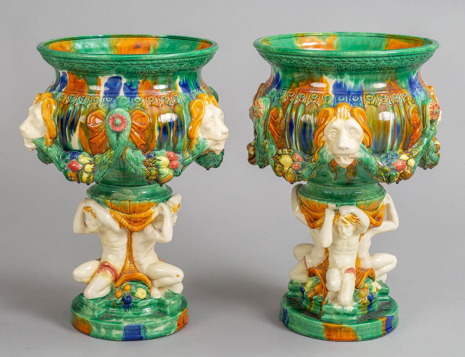 Pair of Majolica multicolored pedestal jardinières, the bowl decorated with bow tied swags of fruit and foliage terminating at lions heads, supported atop a base molded with three partially draped male figures holding up the bowl. Between each of