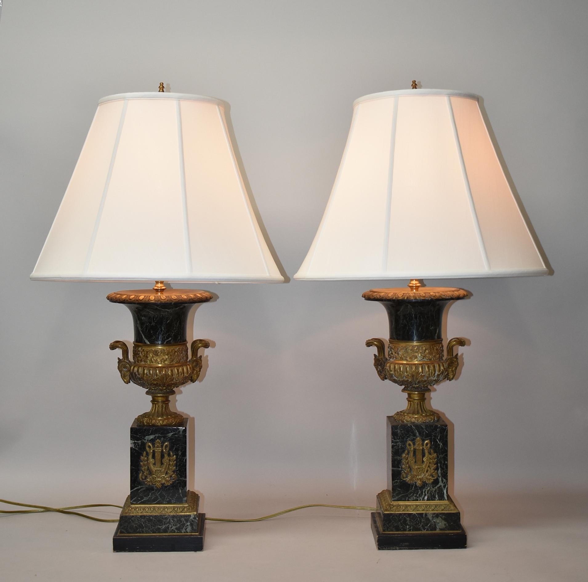 Pair of French Empire style single socket table lamps. Urn shape base with applied figural ram head handles on either side. Bronze lute and swan details in the center. Across the top is a relief border with a grapevine pattern.