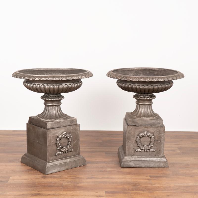 This pair of generous sized cast iron urns resting on a square plinth have been sand-blasted, resulting in an attractive metallic finish. Note the wide and shallow shape of the planter bowl with an egg and dart decorative detail. The plinth features