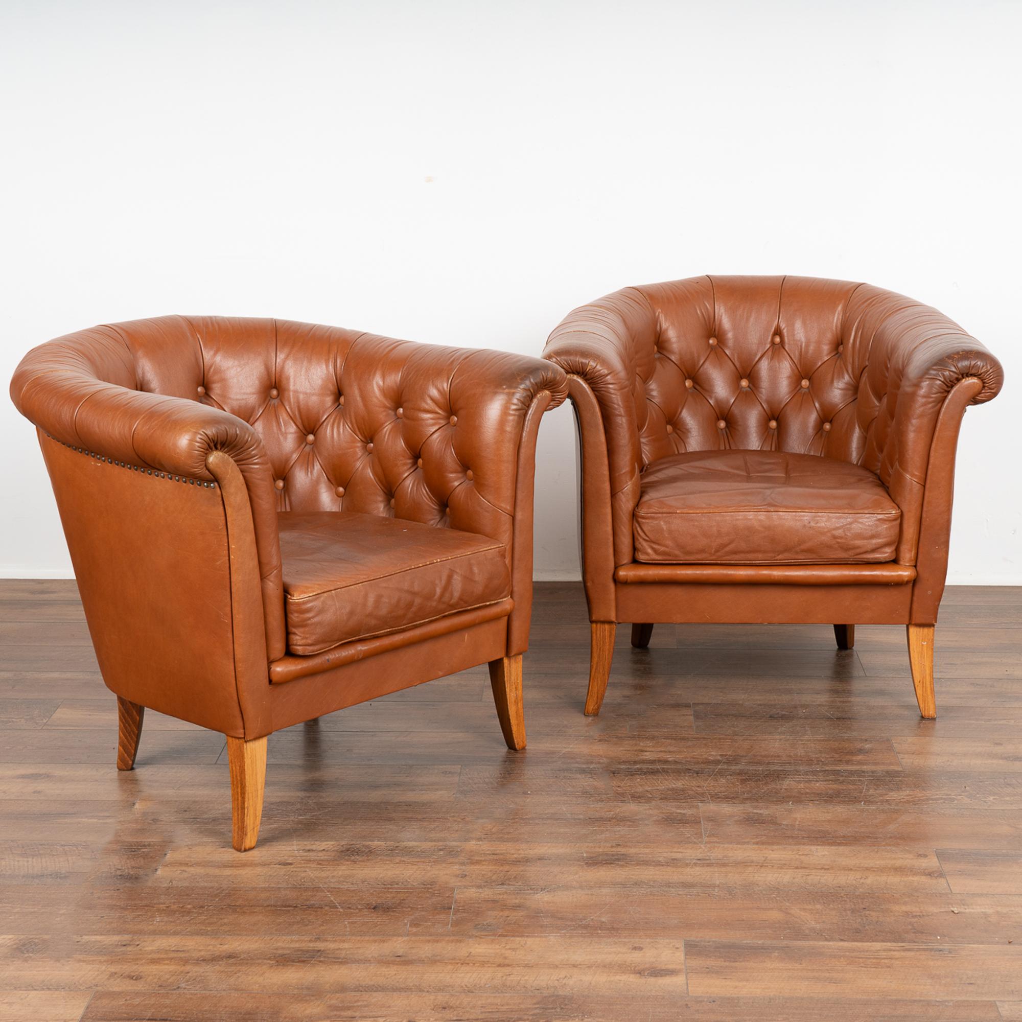 Pair, brown leather barrel back arm chairs with hard wood legs.
Upholstered club chairs in button-tufted cognac leather, slender rolled arms with nail head trim underneath.
Sits low; sold in original vintage condition. Typical age related wear