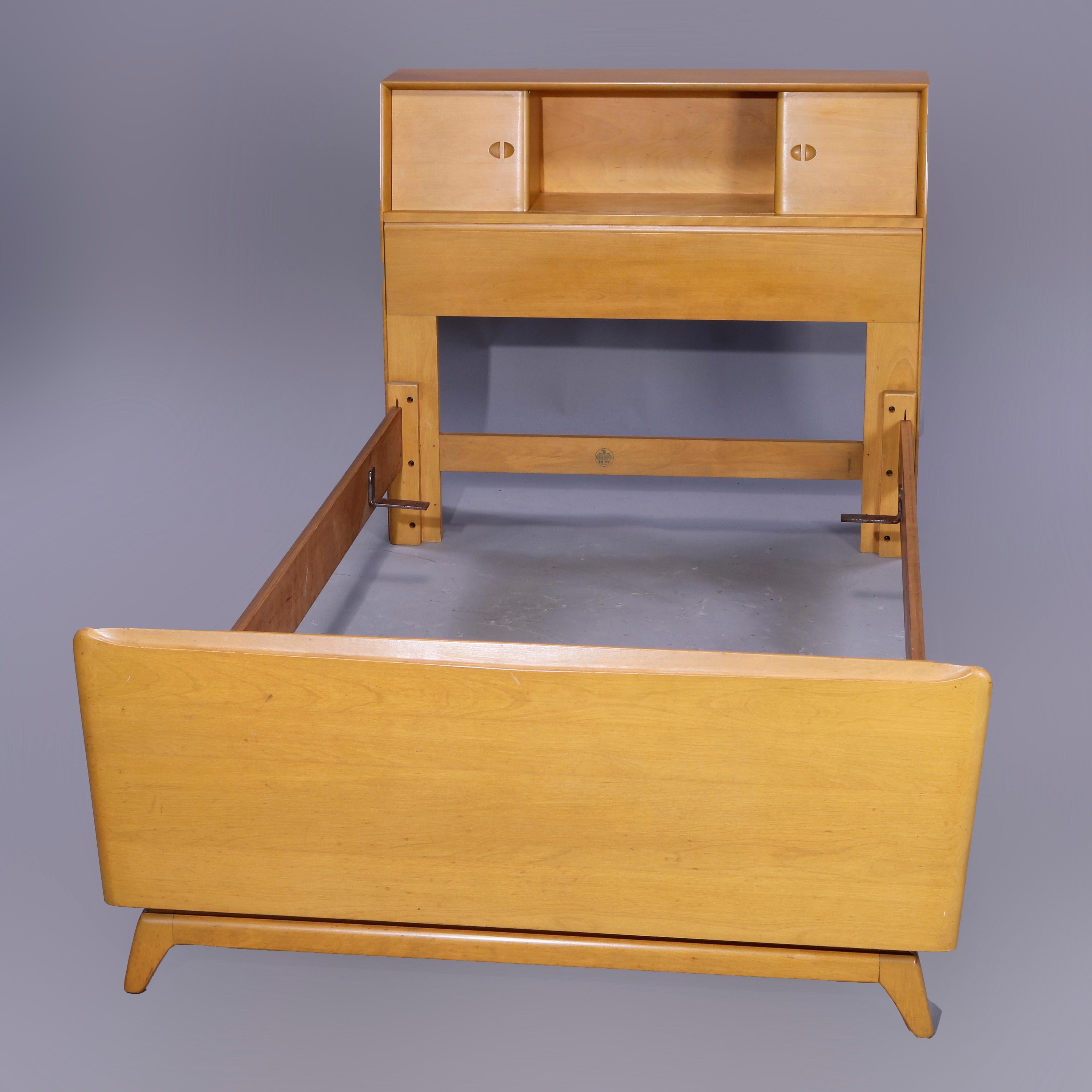A matching pair of Mid-Century Modern twin or single bed frames by Heywood Wakefield offer birch construction with utility headboards having central bookshelf with flanking single sliding door cabinets, raised on splayed legs and having Wheat