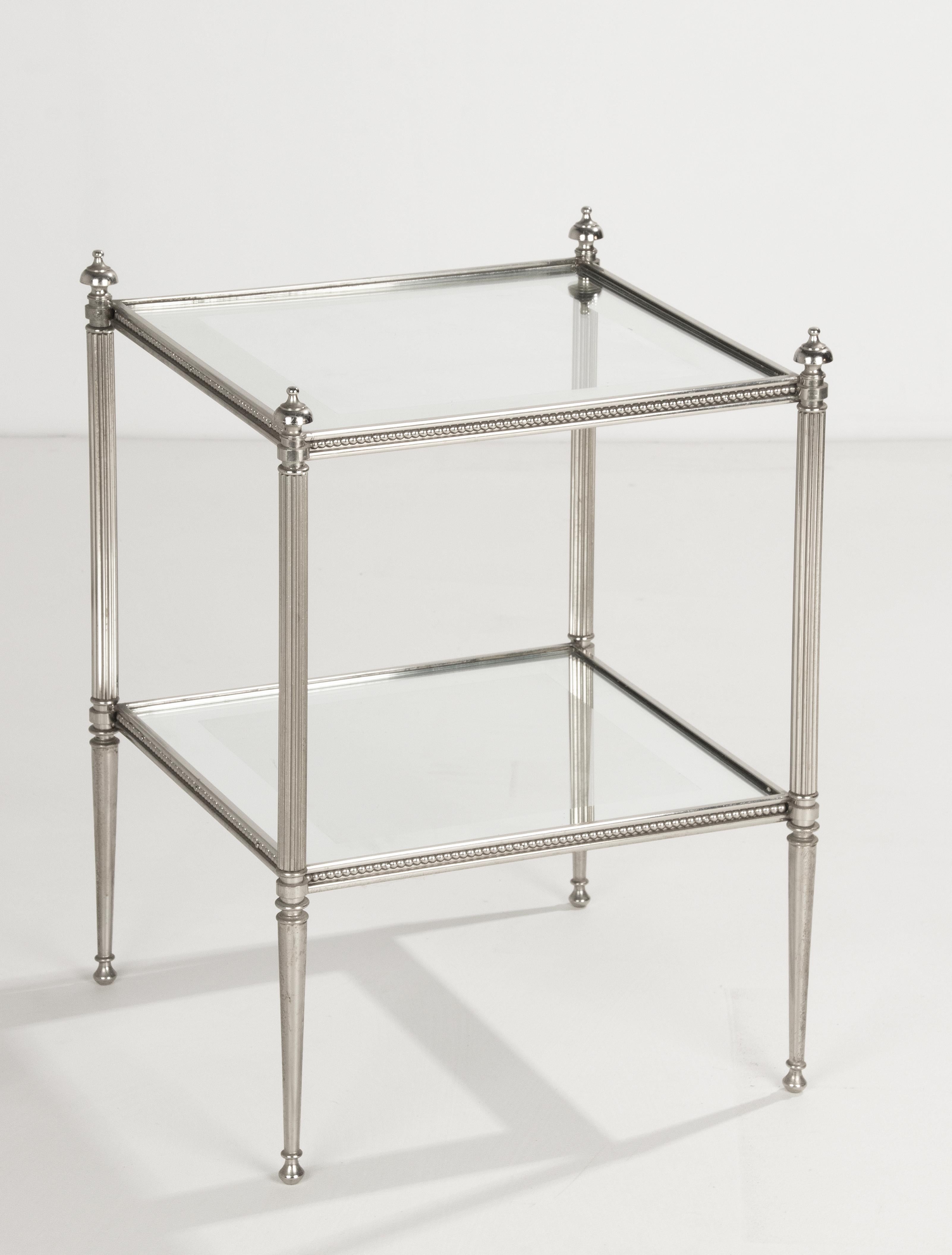 A pair of two-tier side tables made of chromed metal in neo-classical style. In the style of Maison Jansen. It has glass tops with mirrors edges. It's pretty much a pair with a few differences. The beat rim is slightly larger, the feet are slightly