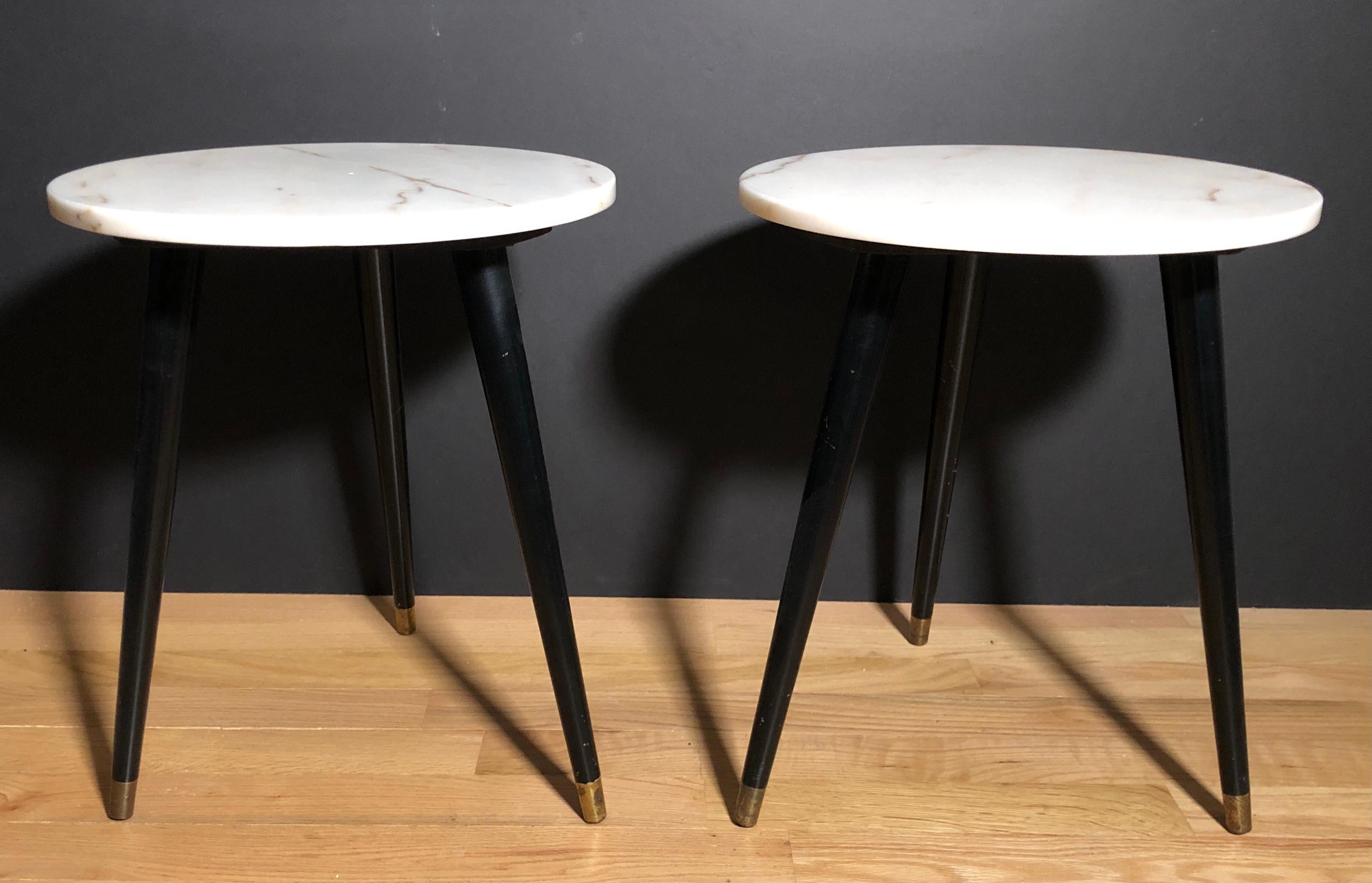 Pair of tripod Mid-Century Modern three leg round marble-top side tables. Off-white marble tops having a slight peach tone with a small amount of veining. Black painted legs with brass caps.
    