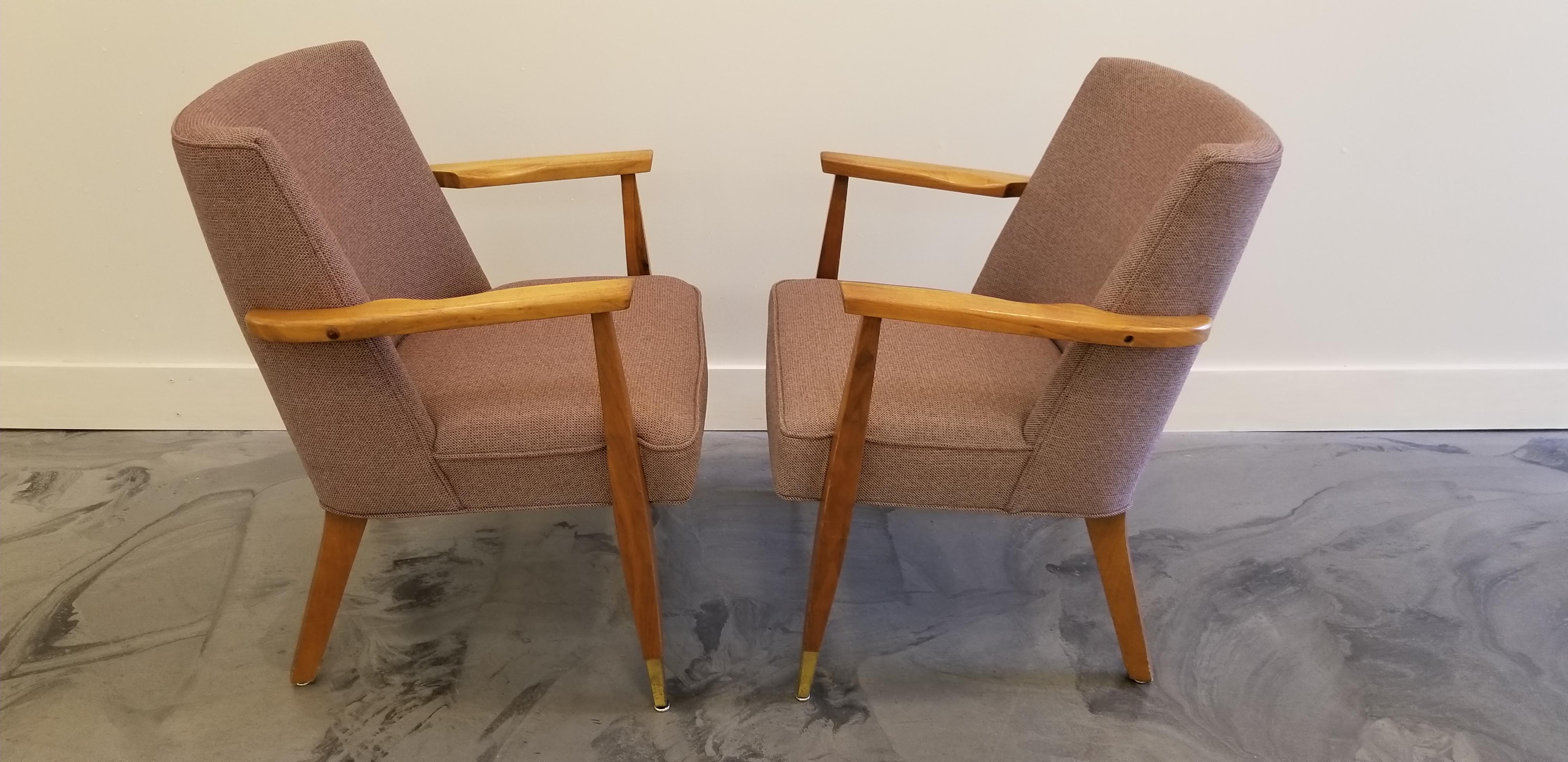 A pair of 1960s armchairs crafted in solid walnut. Unusual hourglass detail to front legs. Classic brass caps to legs. Comfortable, curved backrest. Recent upholstery, but not new.