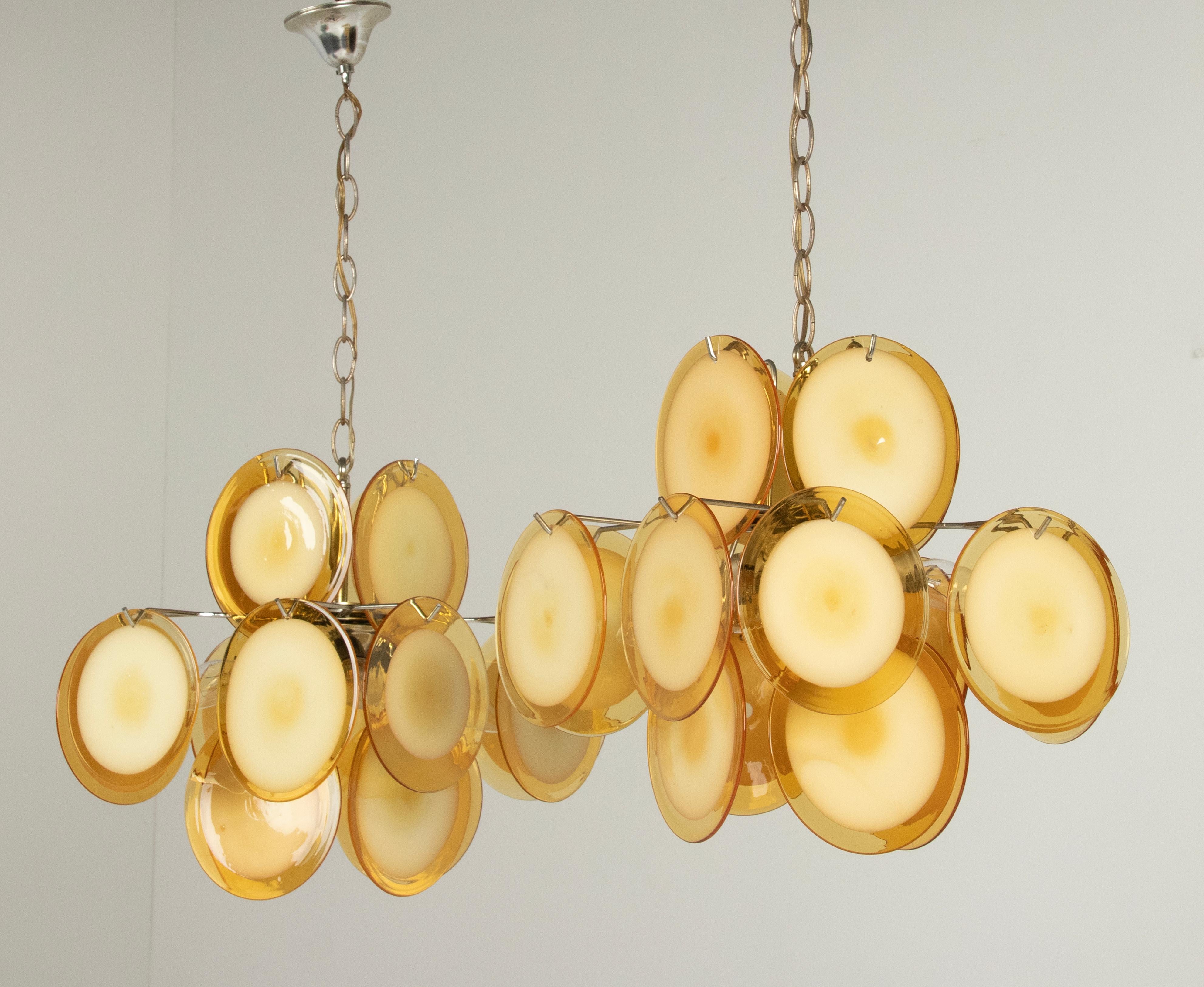 A pair of Italian Murano chandeliers attributed to Vistosi. The chandelier has 16 hand blown glass disks made of yellow/orange Murano glass, hanging in a metal frame. The lamp has 4 lights (E14 sockets), which gives an atmospheric light. Made in