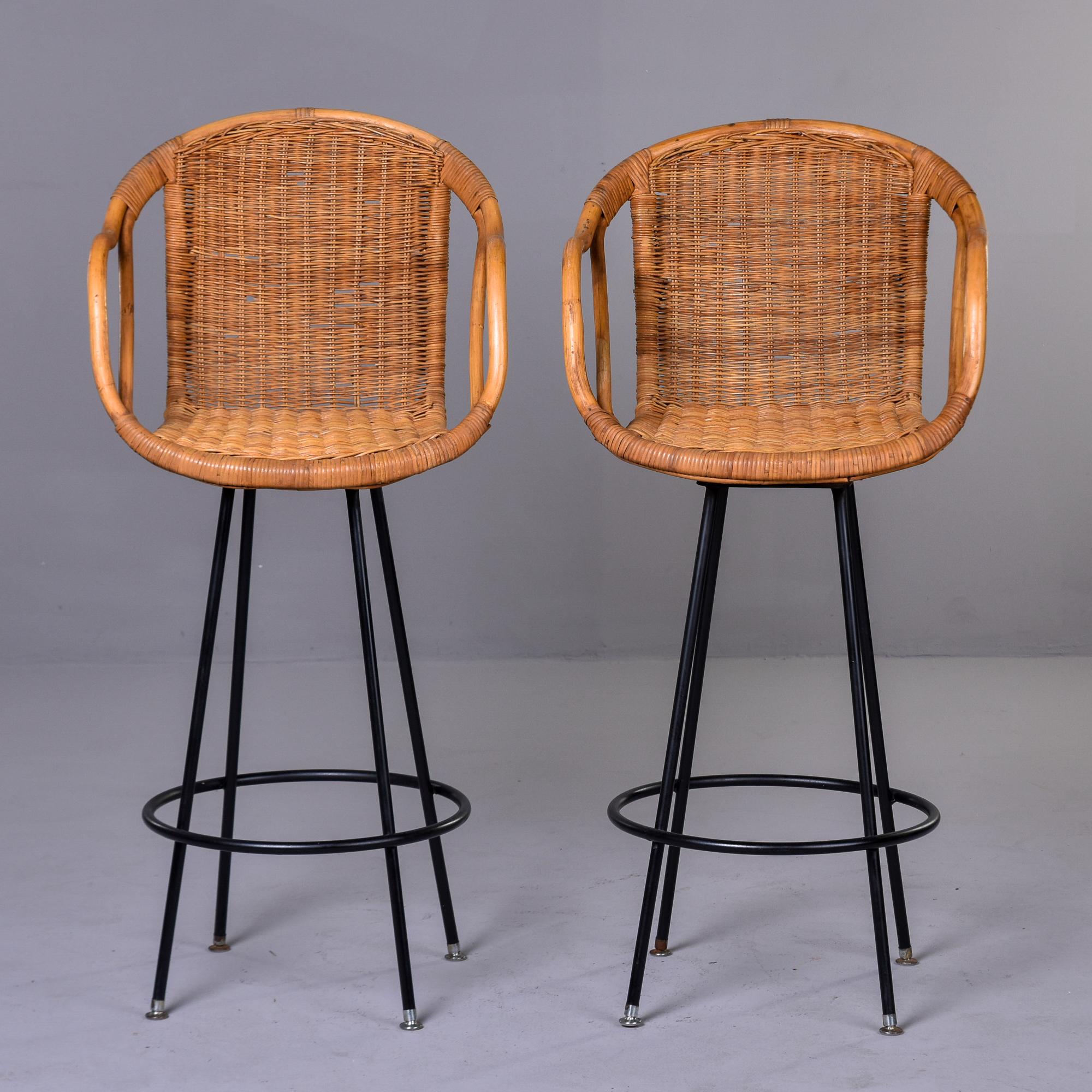 Found in the US, this pair of woven wicker bar height swivel stools date from the early 1960s. Four slender black metal legged base with a round foot rest. Seats are a rounded scoop shape with curved arm rests and cut out sides. Unknown maker. Sold