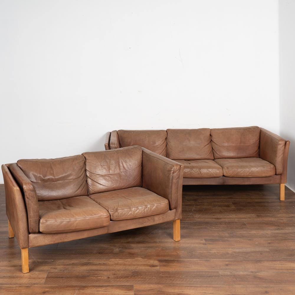 Mid-century modern three-seat sofa and two-seat loveseat set with hard wood birch legs.
Upholstered in vintage brown leather with loose cushions on the sides, seat and back. Each has a zippered cover so cushion inserts are removeable. 
Dimensions of