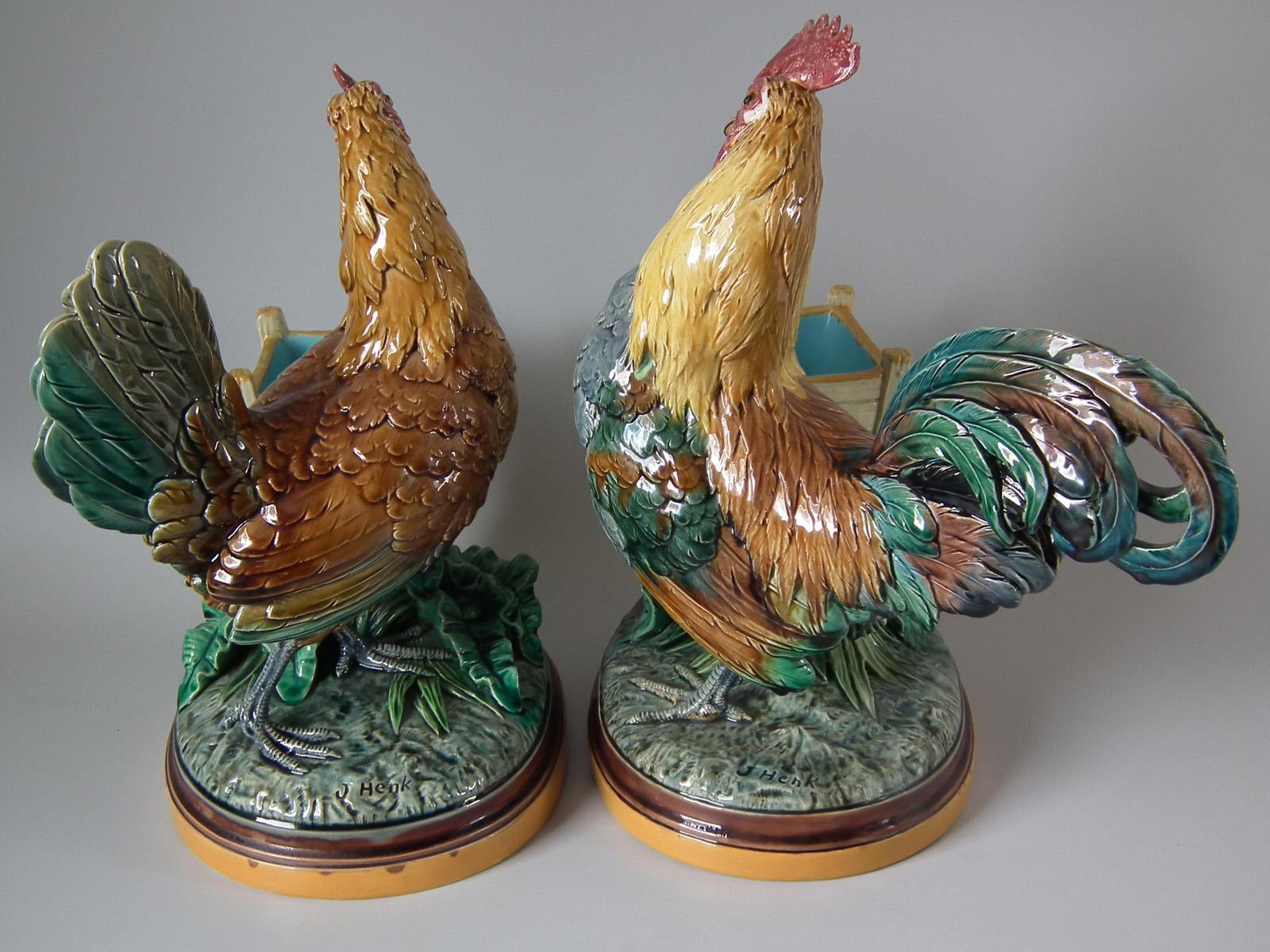 Pair of Minton Majolica vases which feature a hen and rooster stood next to a wooden slated trough. Signed, 'J Henk' to the base. Colouration: brown, green, grey, are predominant. The piece bears maker's marks for the Minton pottery. Marks include a