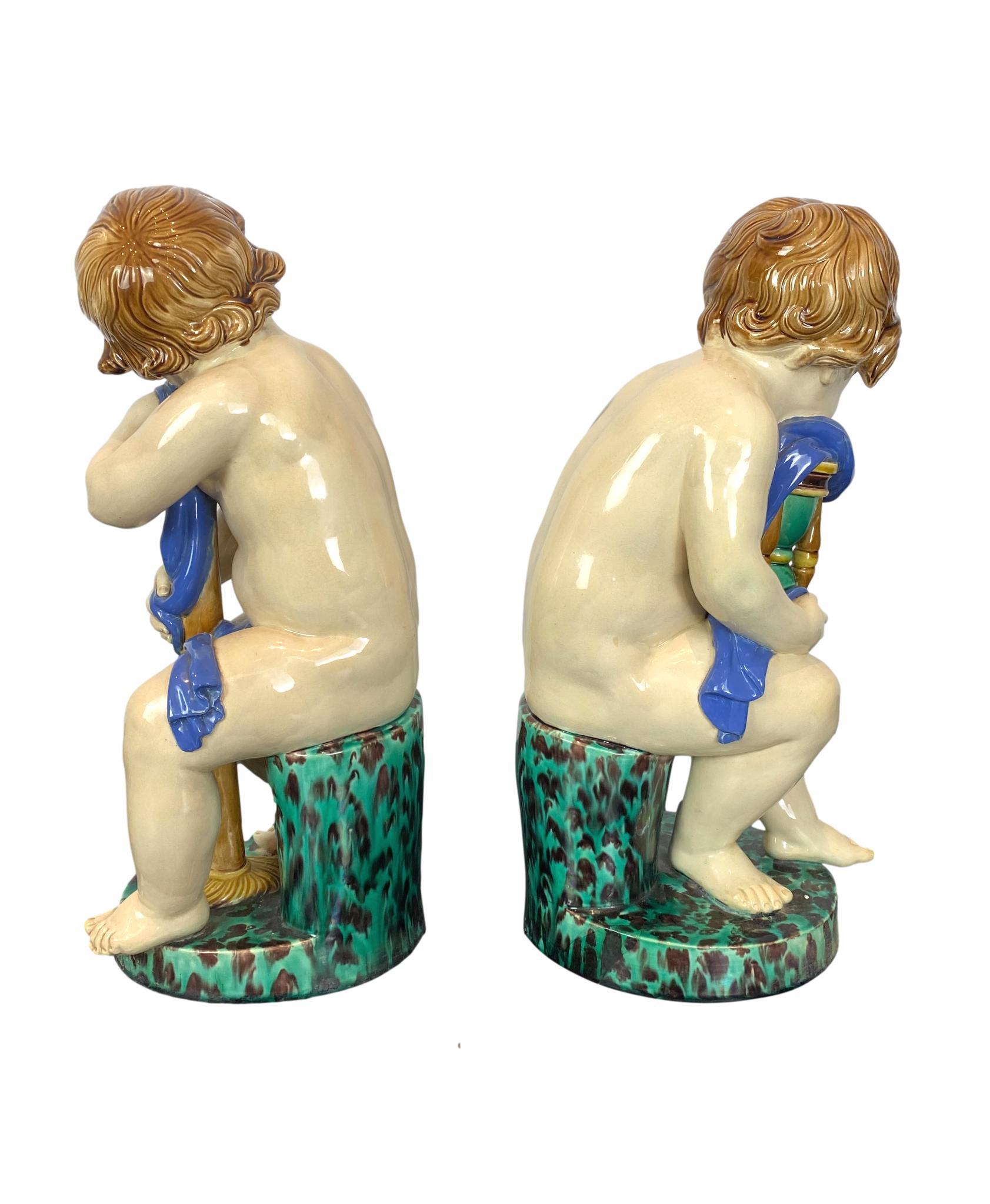 A Pair of Minton Majolica Putti Figures Allegorical of the 'Begining of Time' and 'End of Time,' each seated on a stump and pedestal base glazed in green and brown leopard mottling, one cherub holding an hourglass (Beginning of Time) and the other a