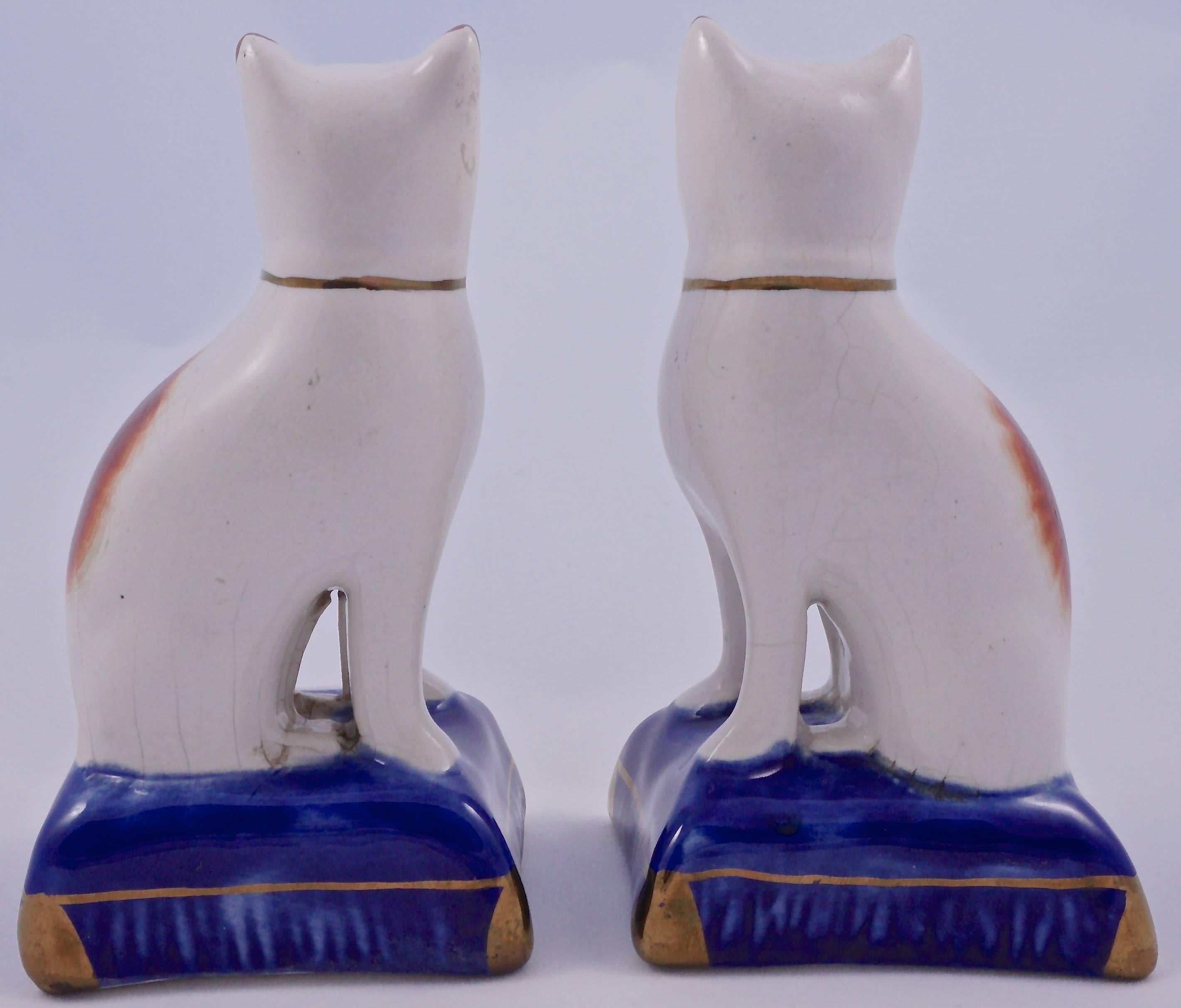 Pair of Staffordshire pottery hand painted cats, which look to be more modern. They are sitting on gold decorated cobalt blue cushions, and have tan decoration with golden bows. There is some minor crazing. The underneath has some very small blue