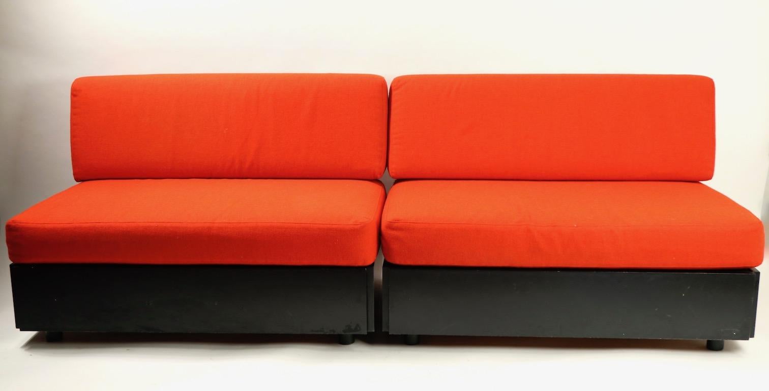 Very nice pair of armless bench form chairs, or sofas, having hidden storage under the seat cushions. The seating units have black finished frames with natural wood veneer based interior storage, and original orange seat cushions (fabric shows minor