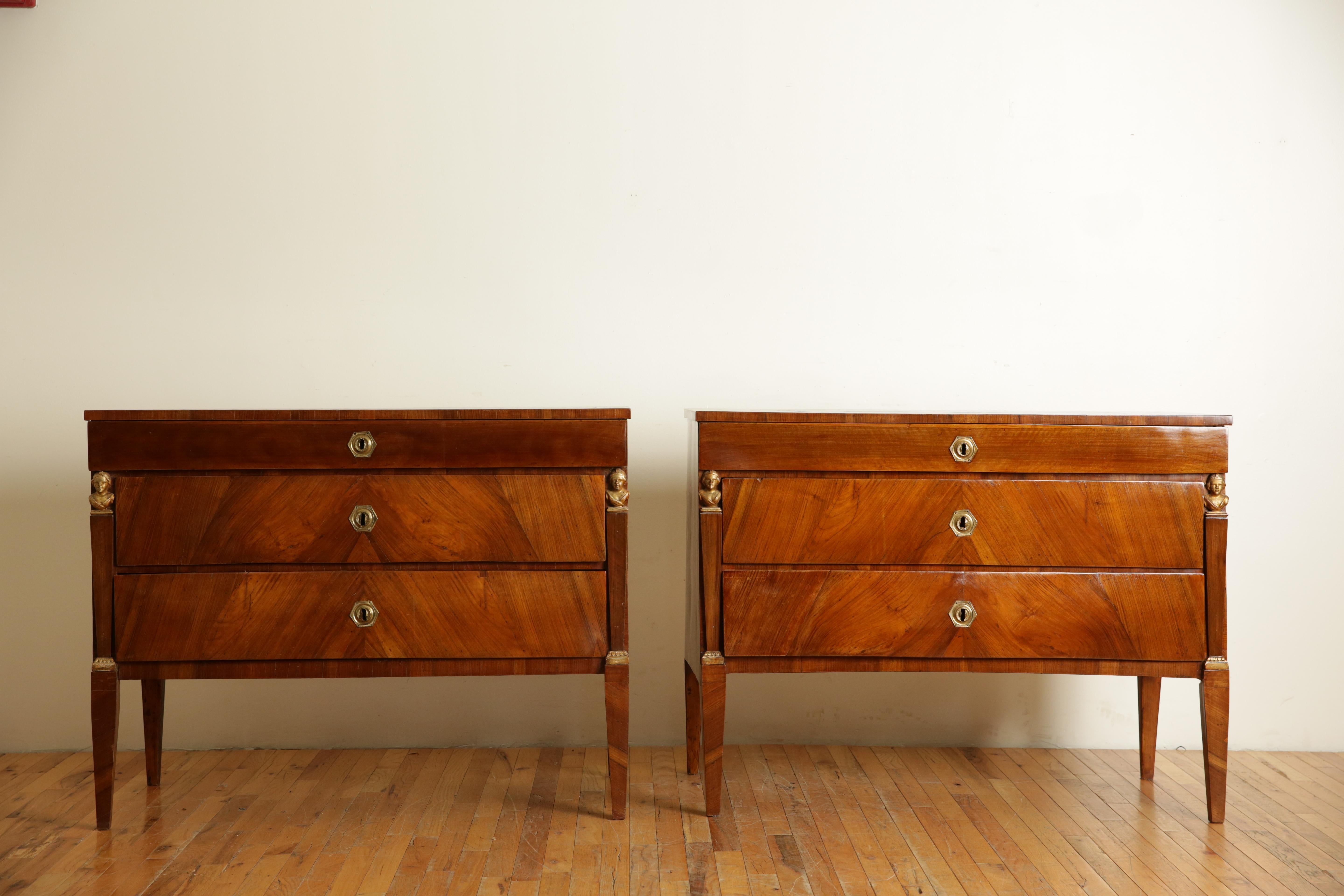 Having rectangular tops with book matched veneers atop conforming cases housing smaller upper drawers above two lower drawers, the drawers flanked by tapering pilasters with giltwood figural bust capitals and carved giltwood bases, raised on square