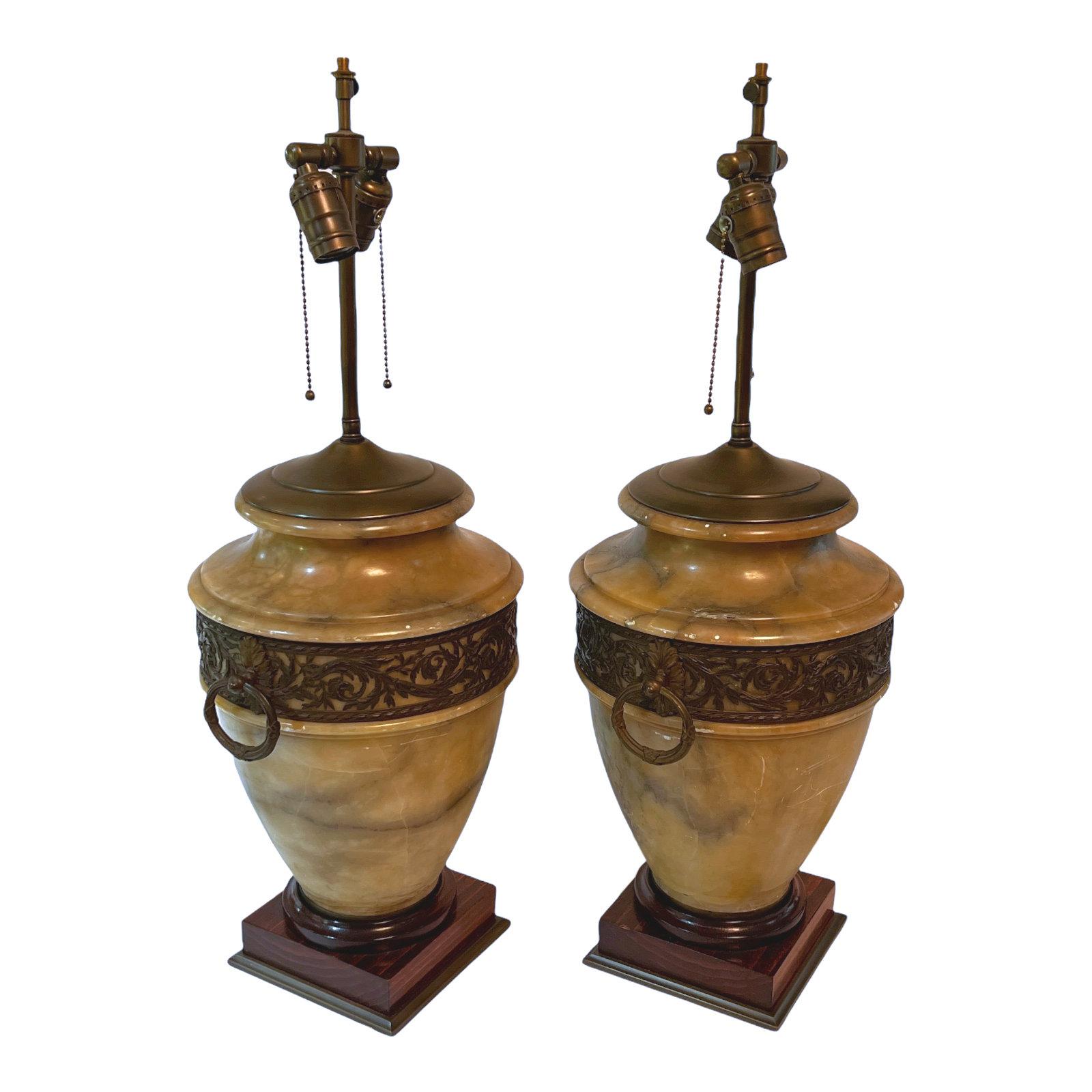 Our pair of table lamps in the neoclassical style have urn forms cut from alabaster stone with brown patinated metal inserts at the shoulder featuring scrolling foliage and ring shaped handles, set upon mahogany pedestals. With sockets and wiring,