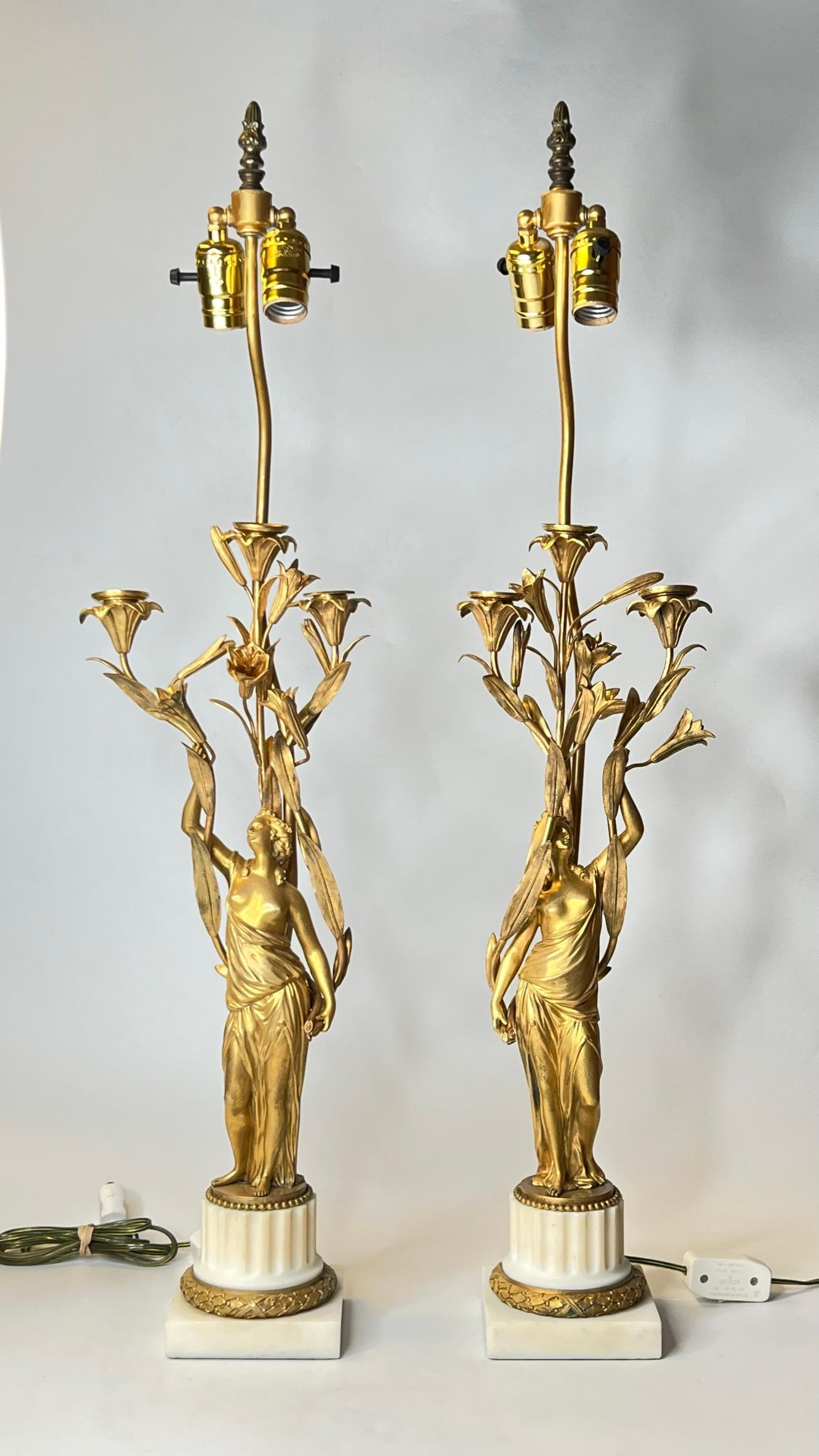 Our pair of nineteenth century gilt bronze and white marble candelabra feature the semi-nude figures of maidens in robes standing among bull rush, and three candle cups emanating from the reeds, with later brass hardware and sockets added. With