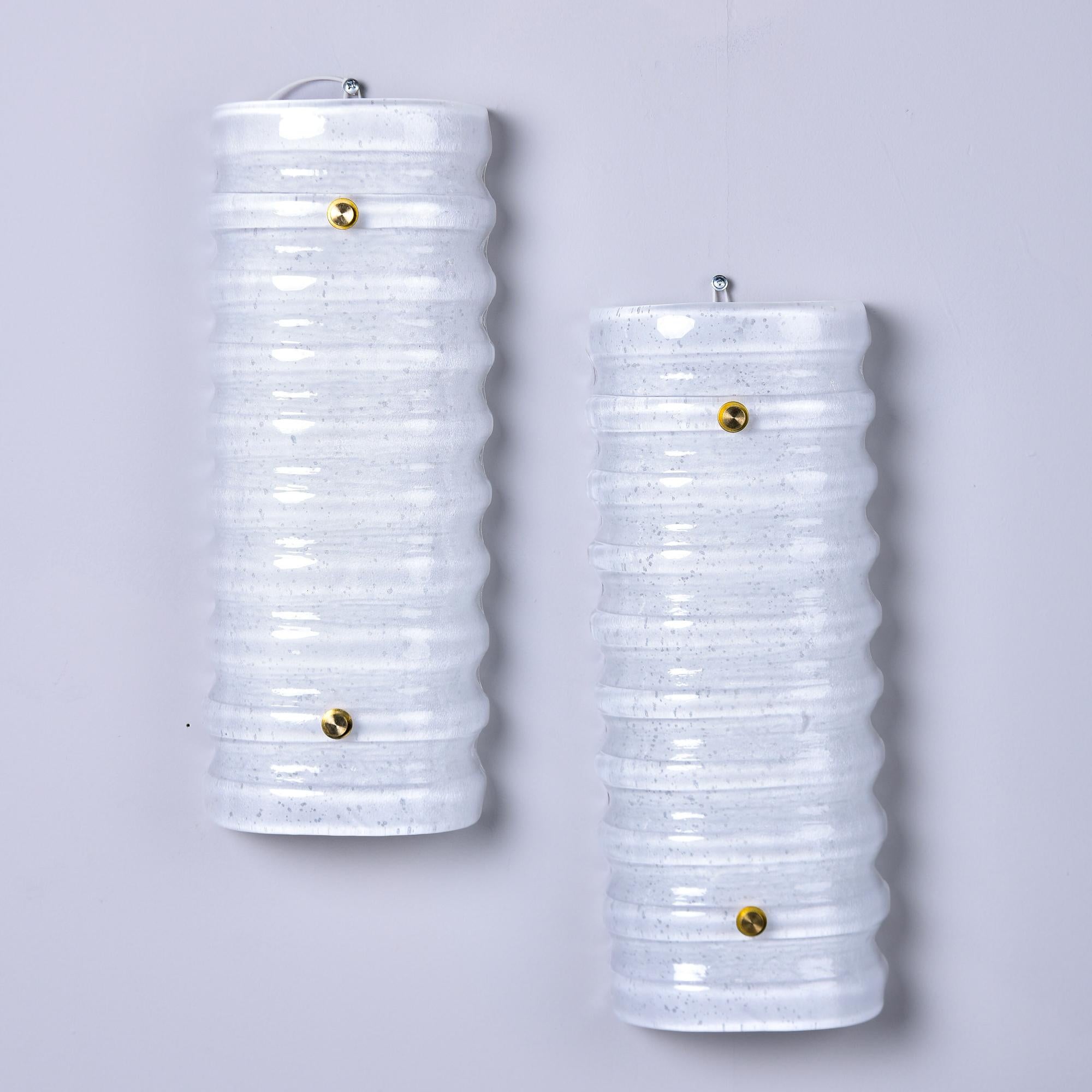 Dieses neue, in Italien hergestellte Paar von Wandleuchtern ist mit Schirmen aus Murano-Glas in opakem Weiß mit gerippter Textur ausgestattet. Messingbeschläge und zwei Steckdosen hinter jedem Lampenschirm. Verkauft und bepreist als ein Paar.