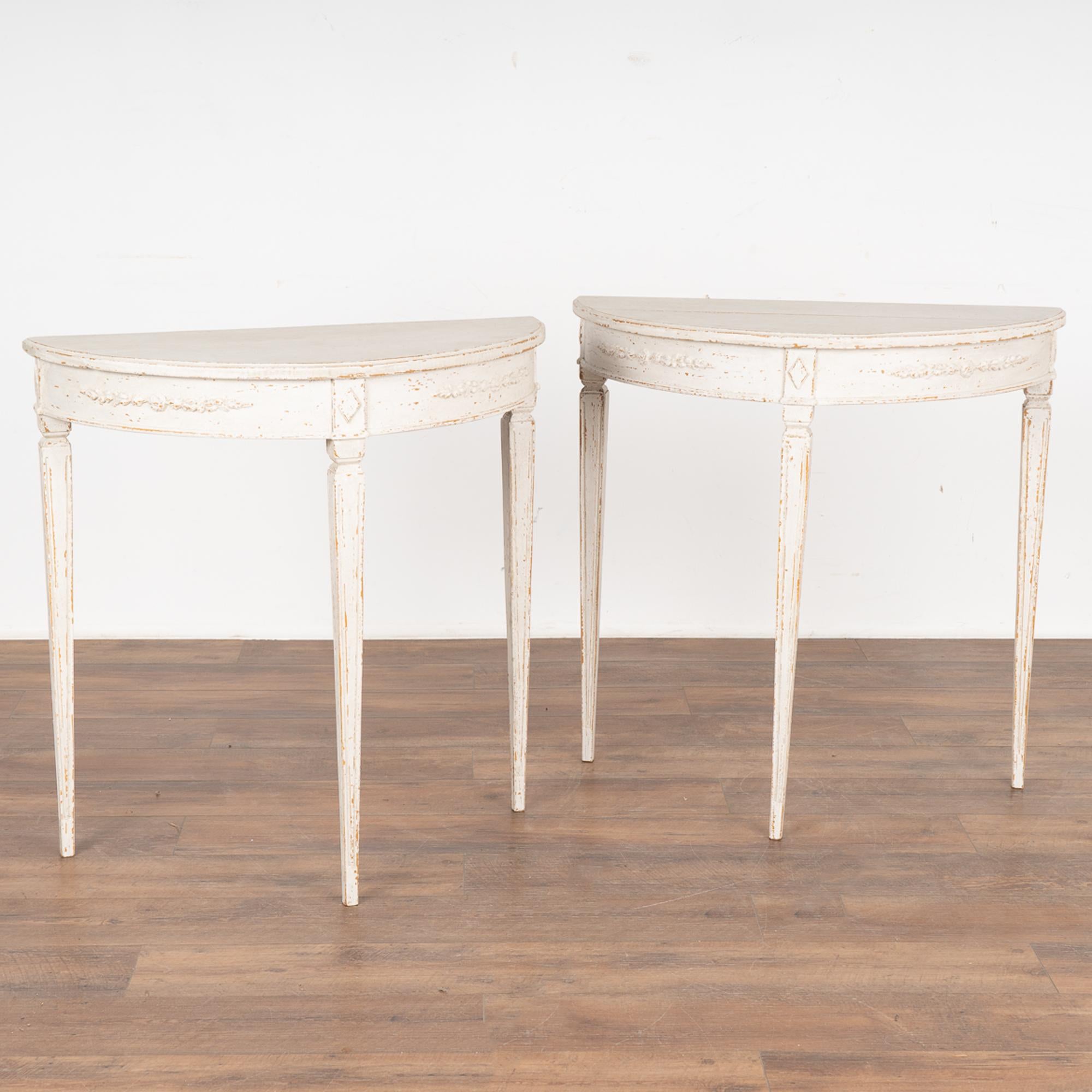 Pair, Gustavian Style demilune side tables or consoles. 
Gently tapered long fluted legs topped by diamond medallion with decorative applied carving along skirt.
Newer professionally applied layered white painted finish, slightly distressed to fit