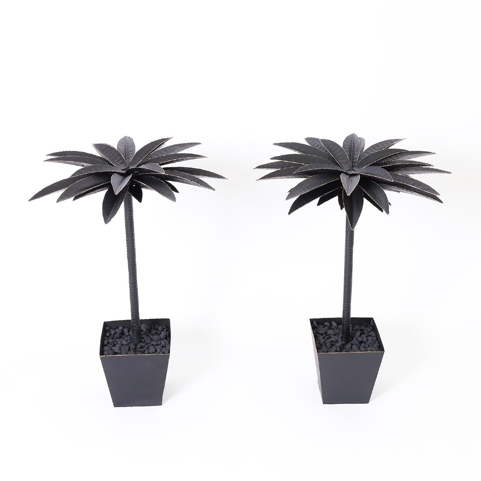 Pair of tole or painted metal potted palm trees with stylized palm leaves having serrated edges on a trunk in a geometric planter with affixed black rocks.