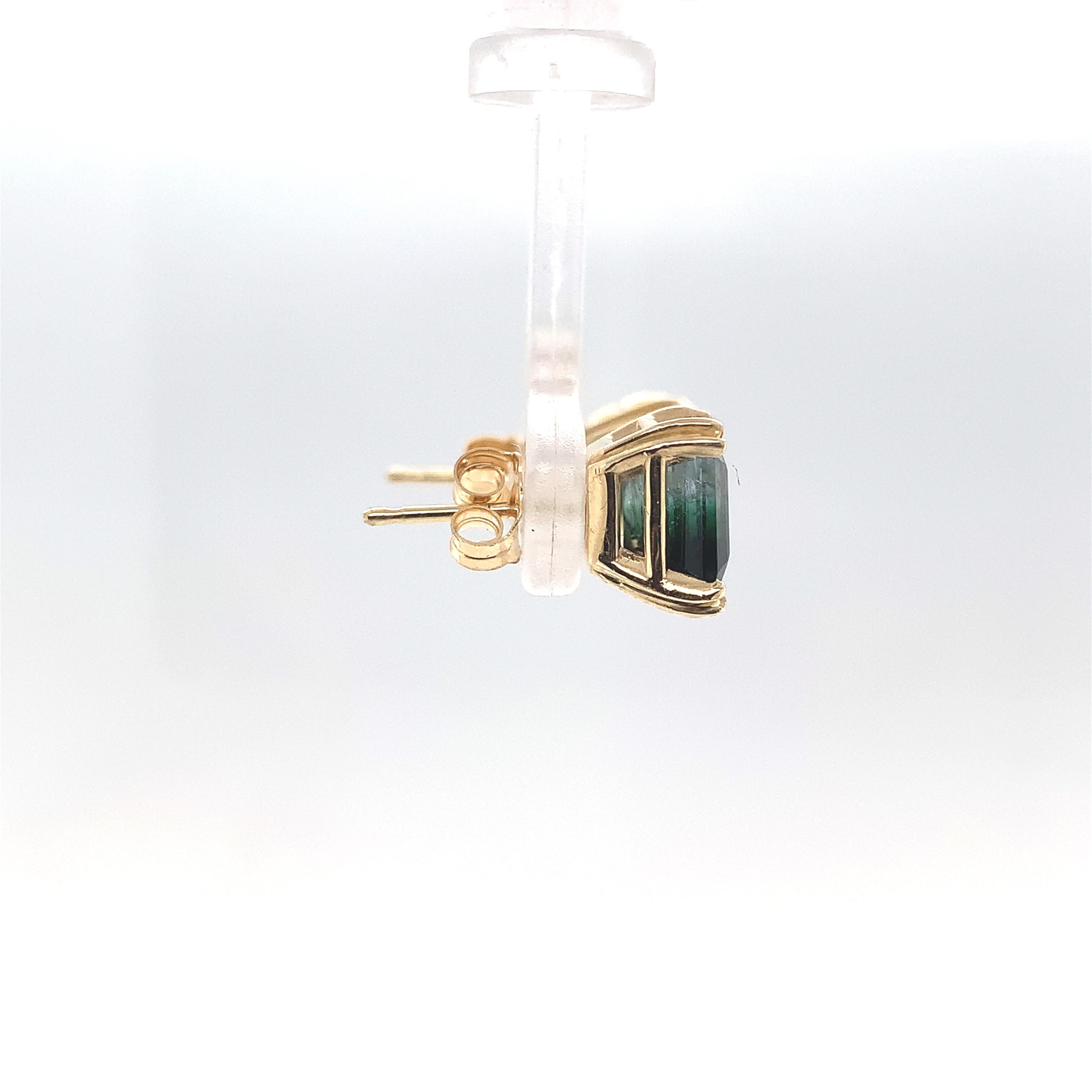 A pair of 14K yellow gold stud earrings featuring a pair of bi-color tourmalines. The tourmalines have color from clear to green to very dark green, almost black. The tourmalines are square step cut and measure about 7.3mm. The pair of tourmalines