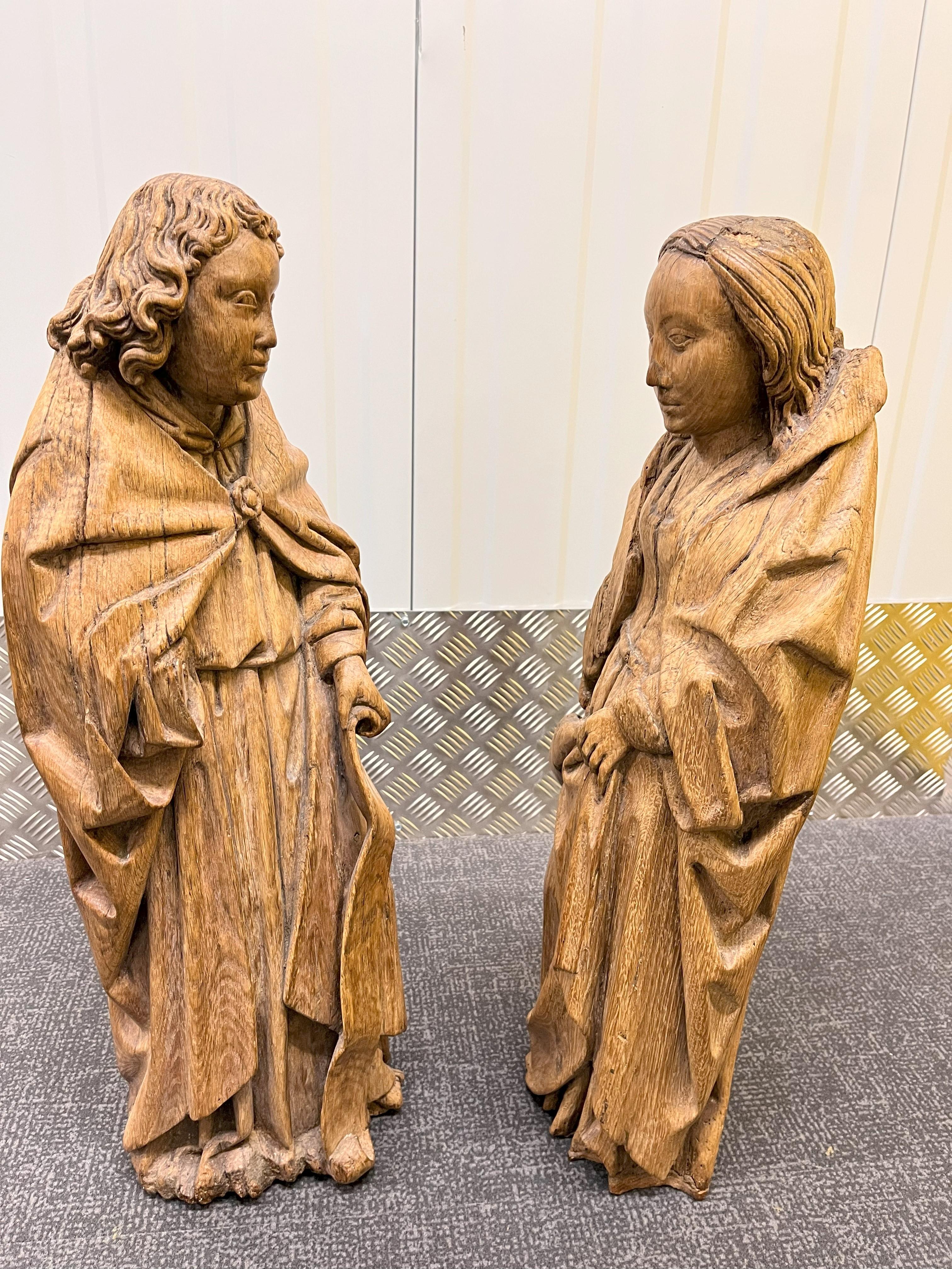 A Pair of 16th Century Oak Saints: One being a male depicted wearing a long hooded cloak held together with a brooch over a flowing tunic tied at the waist and holding a banner in one hand, 28 inches (71 cm) in height, 10 inches wide. The other a