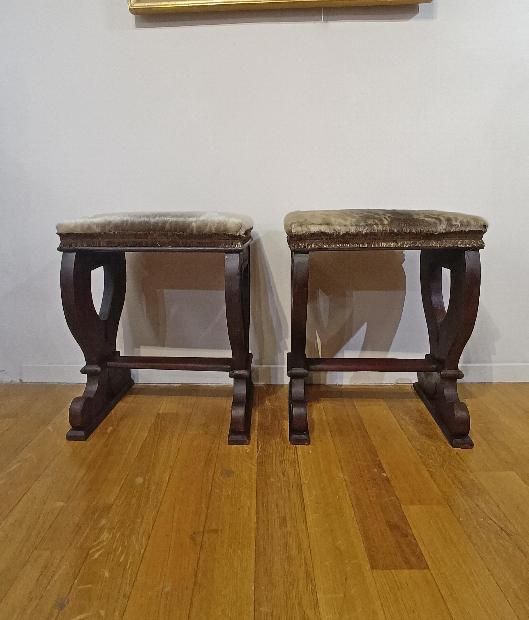 Elegant pair of faldstools in solid hand-carved walnut. The stools are distinguished by their ace-of-cups-shaped legs, joined by linear crosspieces. The design is completed by real fur padding and antique trimmings, giving them a refined and classy