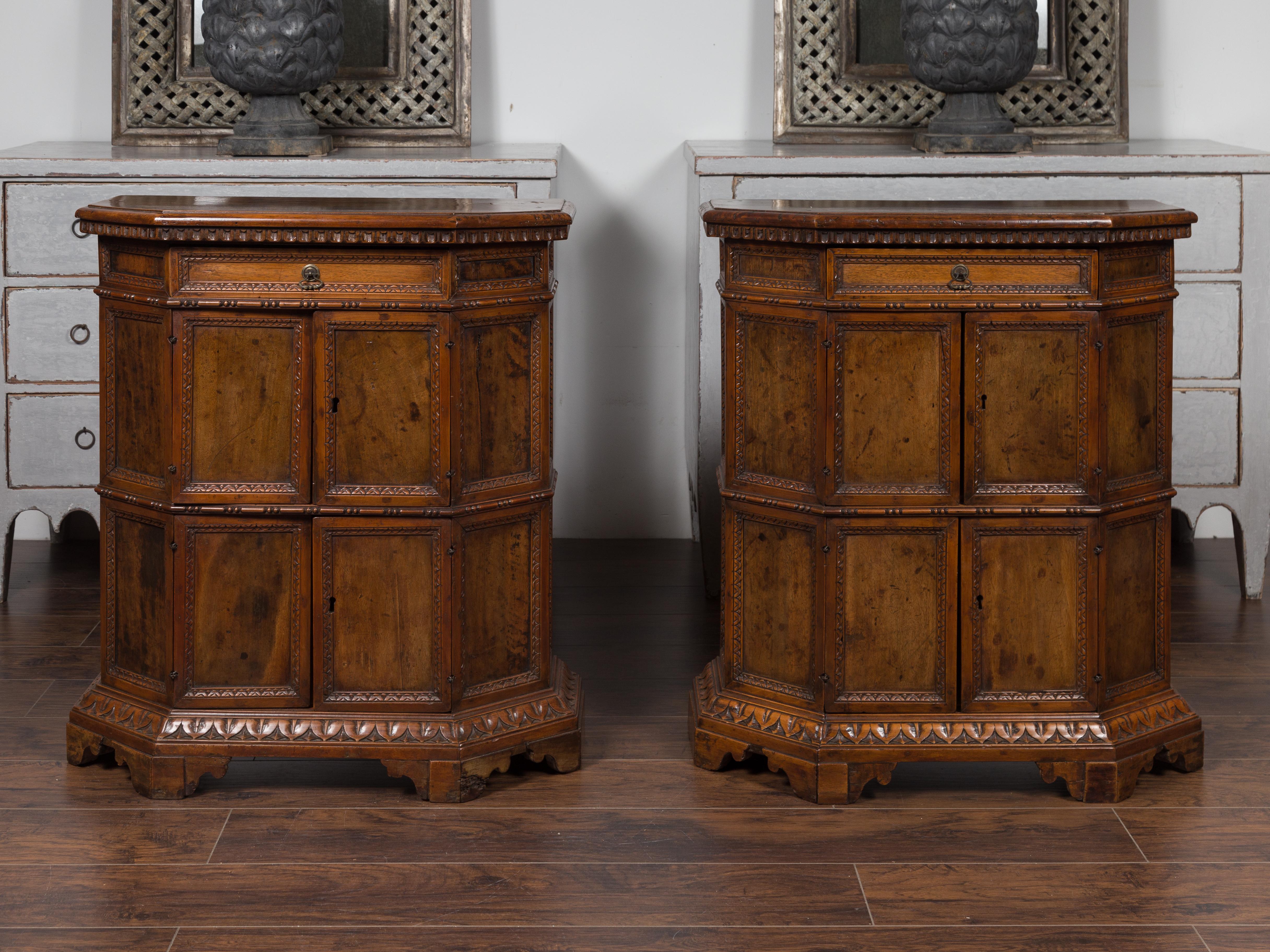 A pair of Italian walnut cabinets from the early 19th century, with canted sides, drawers, doors and carved motifs. Born in Italy during the early years of the 19th century, each of this pair of cabinets features a polygonal top sitting above a