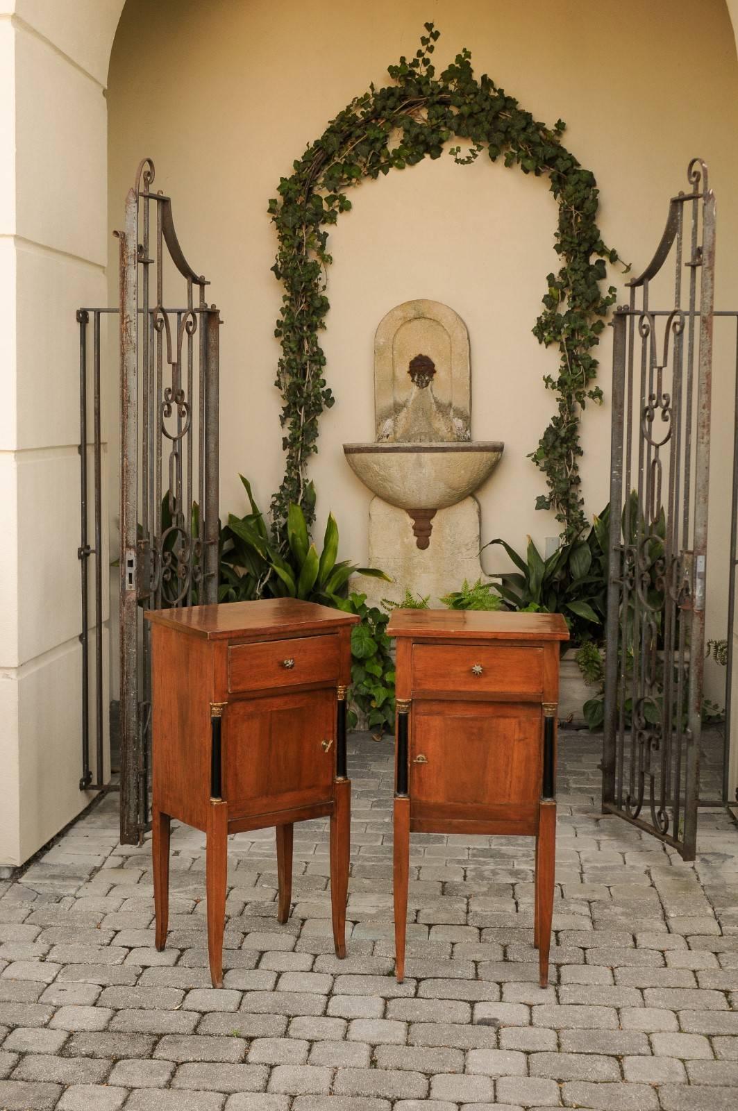 A pair of period Biedermeier walnut stands from the mid-19th century, with single drawer and door, ebonized wood columns and gilt capitals. Born in the first half of the 19th century, each of this pair of petite Biedermeier commodes features a