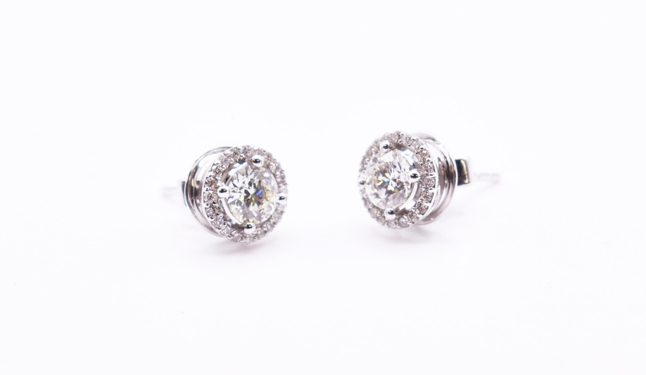 A splendid pair of 18 karat white gold diamond halo stud earrings featuring two bezel set round brilliant cut diamonds of SI1 clarity and I/J colour totaling 1.18 carats, surrounded by 18 prong set diamonds totaling an additional 0.25 carats. The