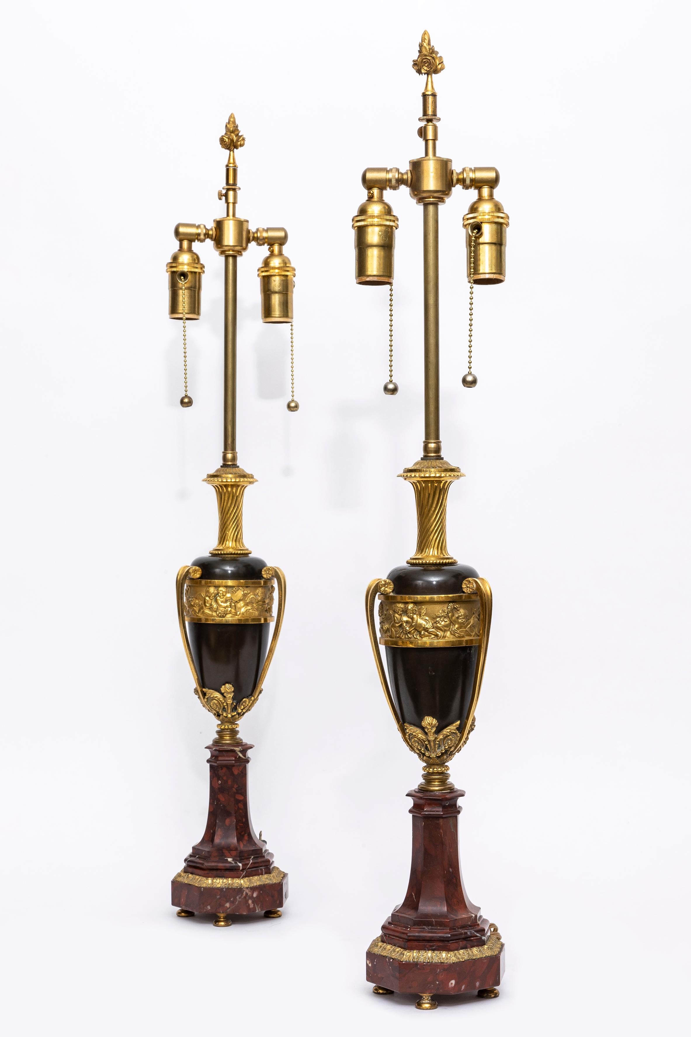  Une incroyable paire de vases en bronze patiné et doré d'époque Louis XVI, transformés en lampes sur des bases en marbre rouge.  
Les lampes ont un corps en forme d'urne avec une frise complexe en bronze. Le relief montre une scène où des anges en