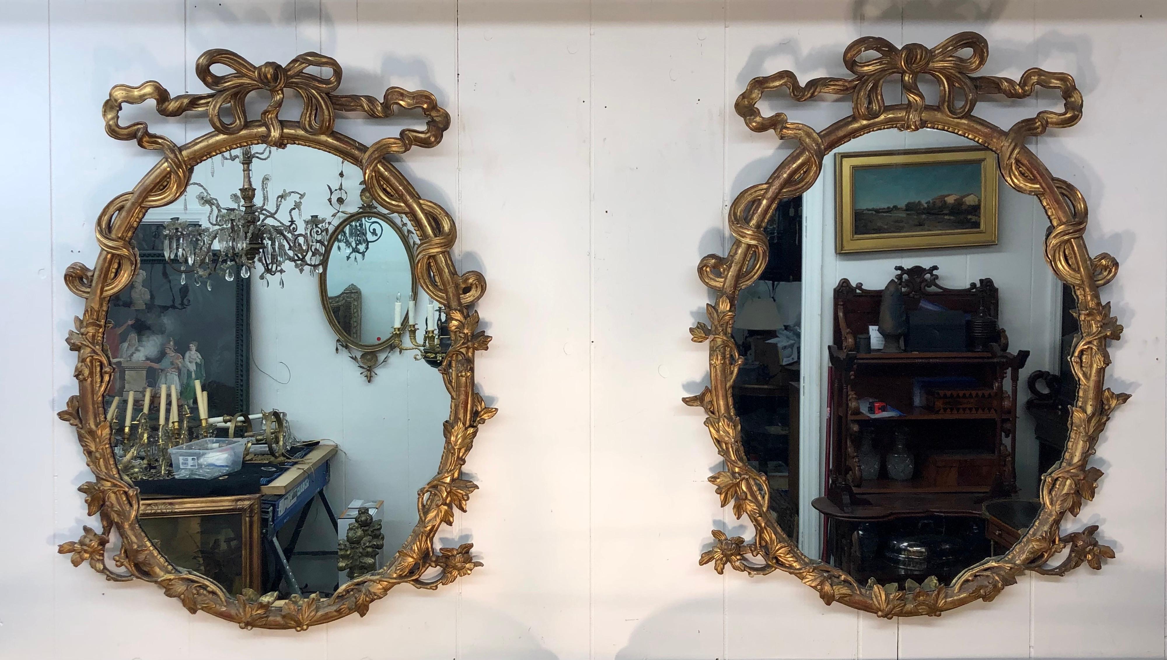 A pair of 18th C. George III period Chippendale oval carved giltwood rococo mirrors. The pierced Ribbon Cartouche is above the oval plates within elaborate rocaille frames with pierced ribbon and foliate vine carving. The elegant Rococo mirrors are