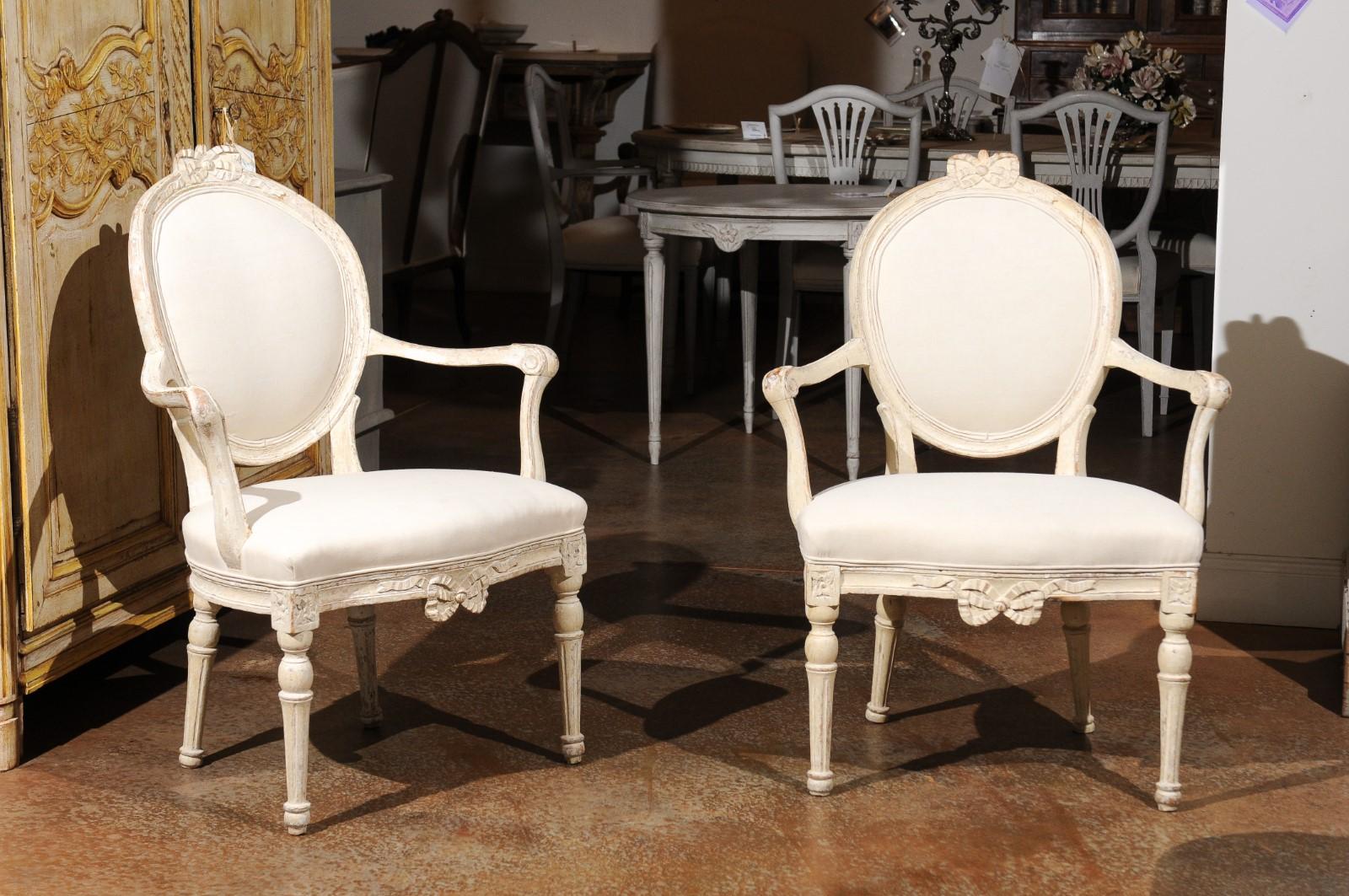 A pair of Danish 18th century Louis XVI painted wood oval back armchairs with bow motifs, scrolled arms and new upholstery. Born in Denmark during the 18th century, each of this pair of wooden armchairs features an oval back accented with a