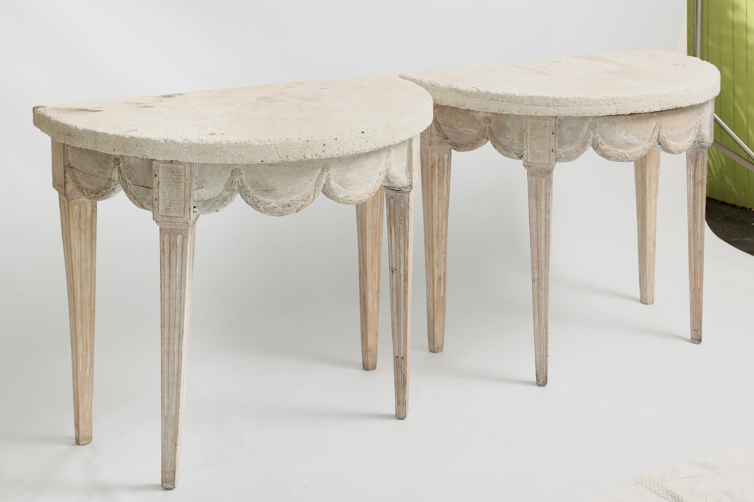 Pair of Demilune console tables, each having tops of old concrete, on painted walnut base, with each apron carved as swagged festooning, raised on square, fluted, tapering legs.

Stock ID: D9288.