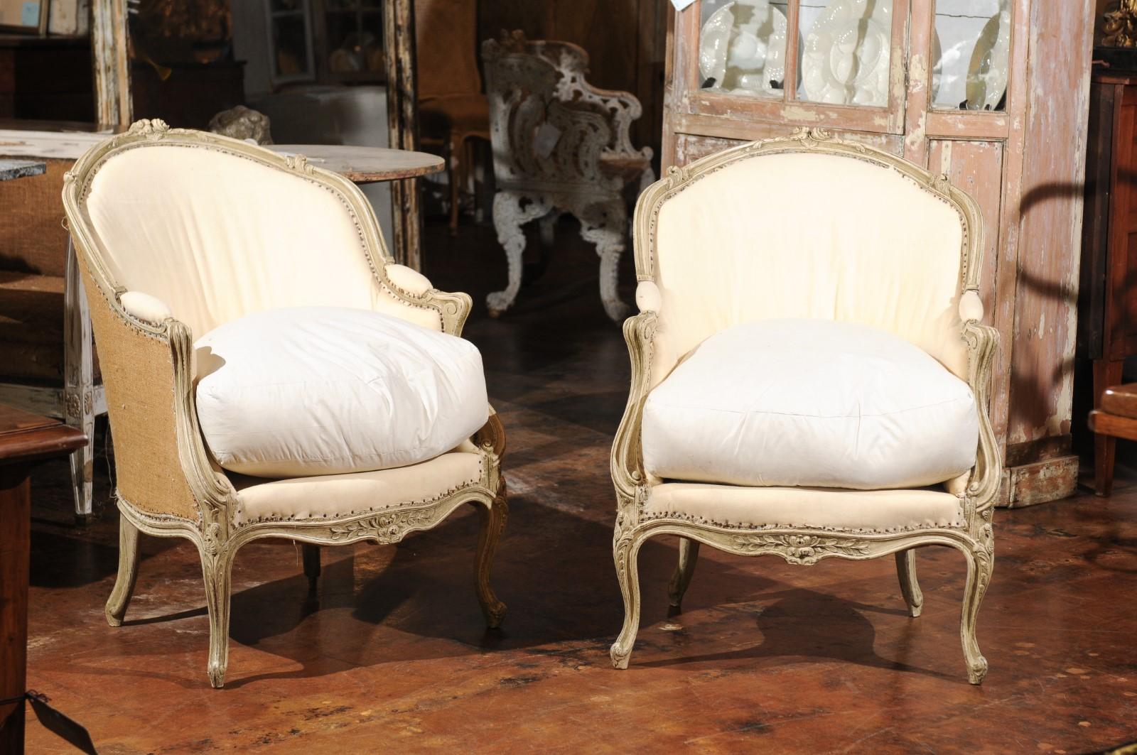 A pair of French Louis XV 18th century painted bergères chairs with wraparound backs and cabriole legs. Each of this pair of French painted bergères features a wraparound back adorned with an exquisitely carved rosette on the crest. This back flows