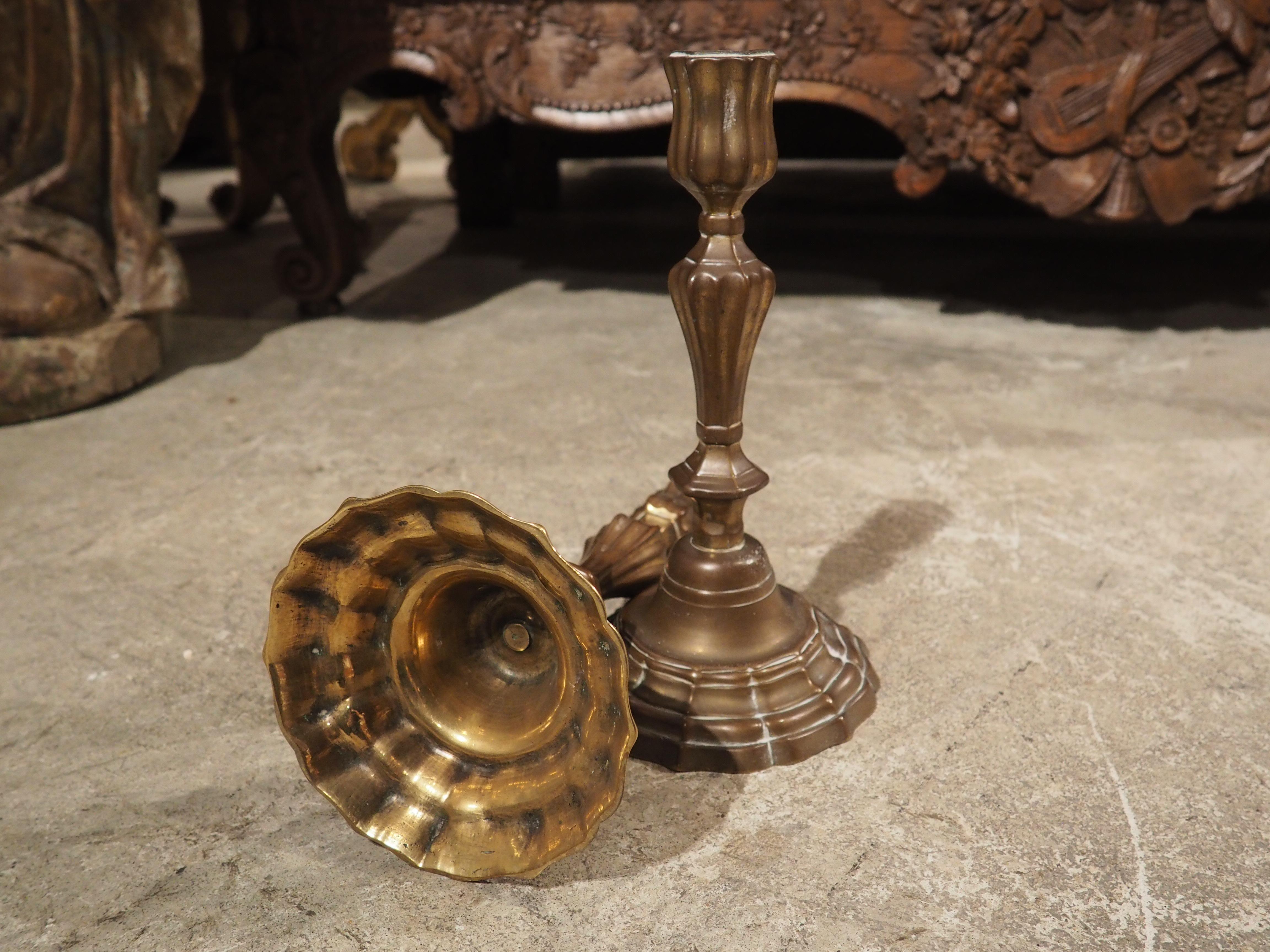 Cast in France during the 1700s, this pair of bronze candlesticks has a lovely patination, giving them a glossy brown color. The highly contoured and curved bases are mirrored by the recessed lobes that embellish the shafts and capitals that are