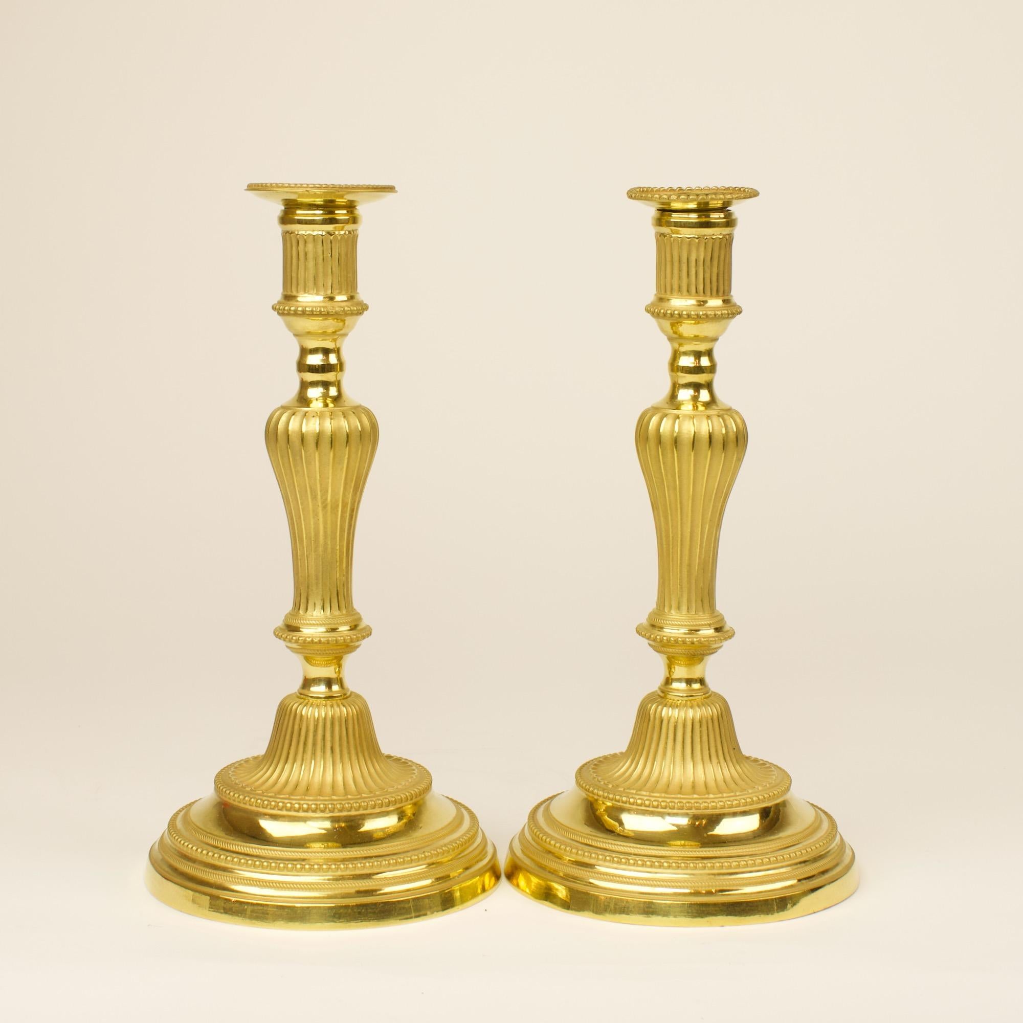 Pair of 18th century French transition Louis XVI gilt bronze candlesticks

A baluster-shaped tapering fluted stem on a large round base and fluted plinth with pearl frieze. A cylindrical fluted nozzle with drip pans (slightly different from each